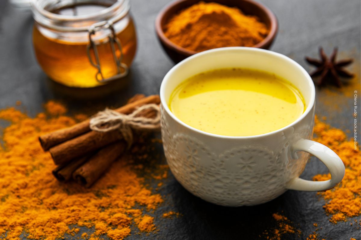 Eine Tasse goldene Milch steht auf einem Tisch. Um sie herum sind ein Glas mit Honig, ein Holzschälchen mit Kurkuma, Zimtstangen und etwas Anis zu sehen. Kurkuma-Pulver ist auf dem Tisch verstreut. Die Tasse ist weiß mit floralem Muster