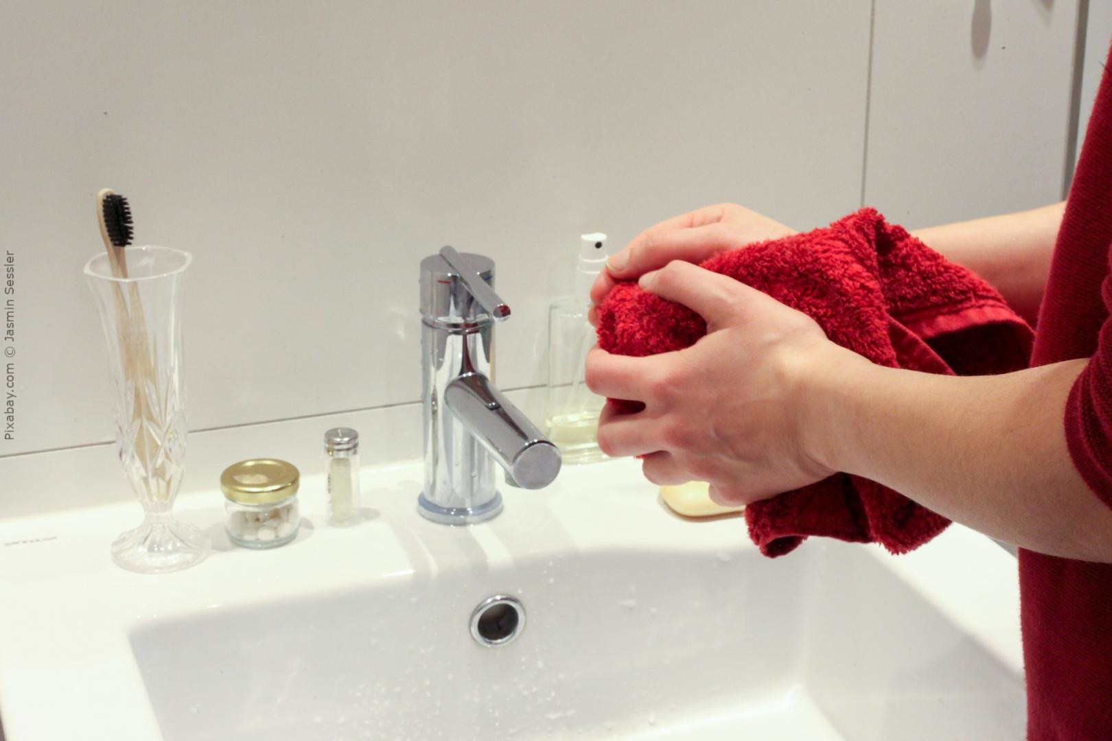 Eine Person steht am Waschbecken und hält ein rotes Handtuch in den Händen. Auf dem Waschbeckenrand befinden sich verschiedene Gegenstände, darunter ein Zahnputzglas mit einer Zahnbürste, eine kleine Dose und eine Flüssigseifenflasche.