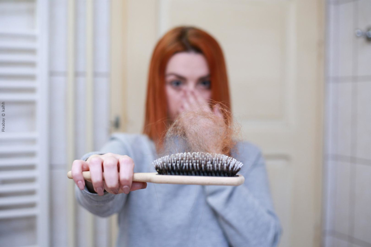 Eine Frau mit langen, roten Haaren hält eine Haarbürste voller ausgefallener Haare. Die Frau schaut besorgt und hält sich die Nase mit der anderen Hand. Der Hintergrund zeigt ein unscharfes Badezimmer mit beige-weißen Wänden und Fliesen.