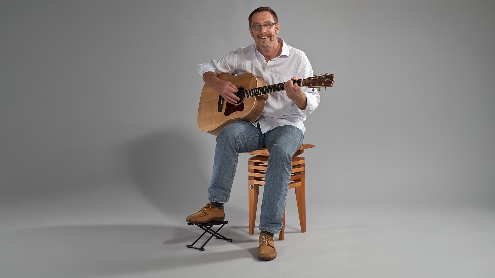 Mann sitzt mit Gitarre auf einem ergonomischen MiShu-Stuhl aus Holz, nutzt eine Fußstütze und spielt entspannt im Studio – Symbol für rückenschonendes, bewegtes Sitzen beim Musizieren mit natürlicher Aufrichtung und Komfort.