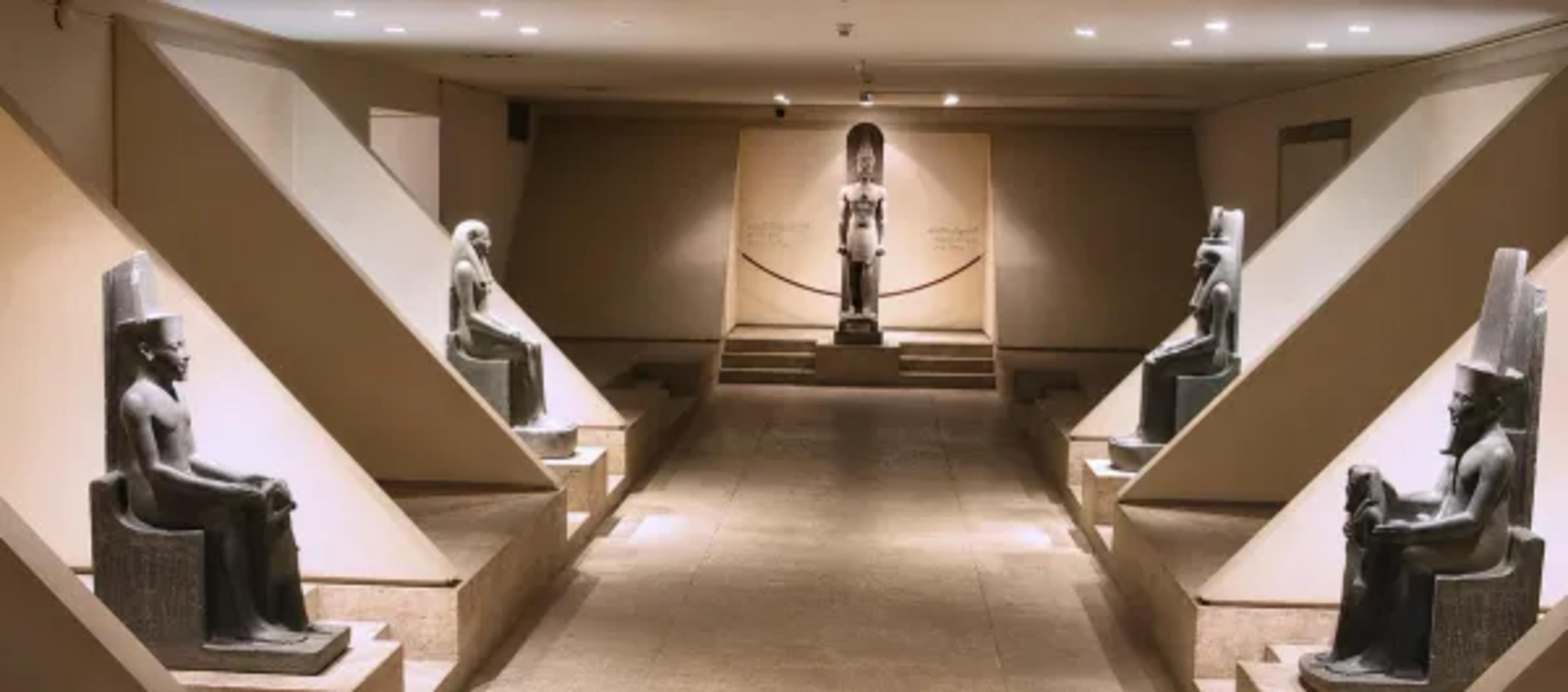 Main hall of the Luxor Museum, with seated and standing black-granite royal statues — including a tall standing Amenhotep III at the far end — displayed on stepped plinths in a minimalist beige gallery.