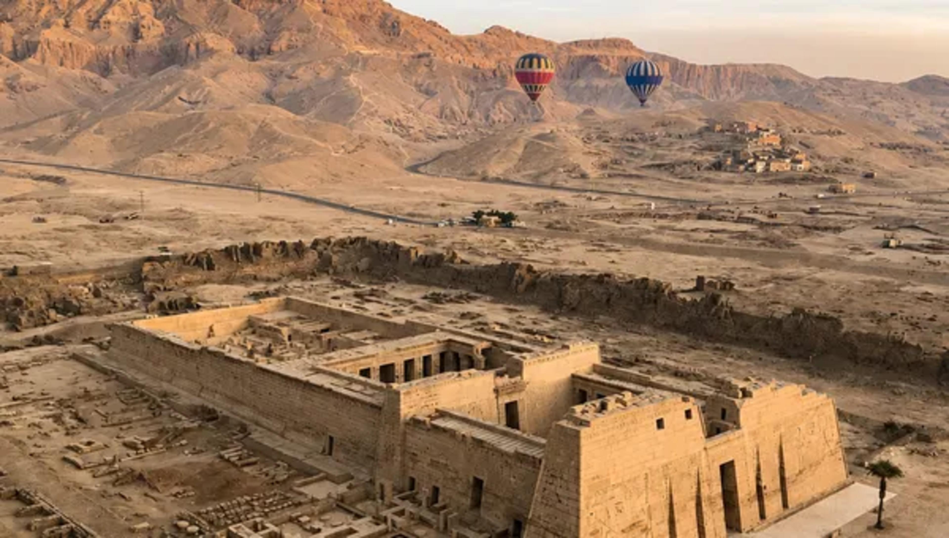 Aerial view of Medinet Habu — the mortuary temple of Ramesses III on the West Bank of Luxor — with its massive pylons and hypostyle hall in the foreground and three hot-air balloons drifting over the Theban hills behind.
