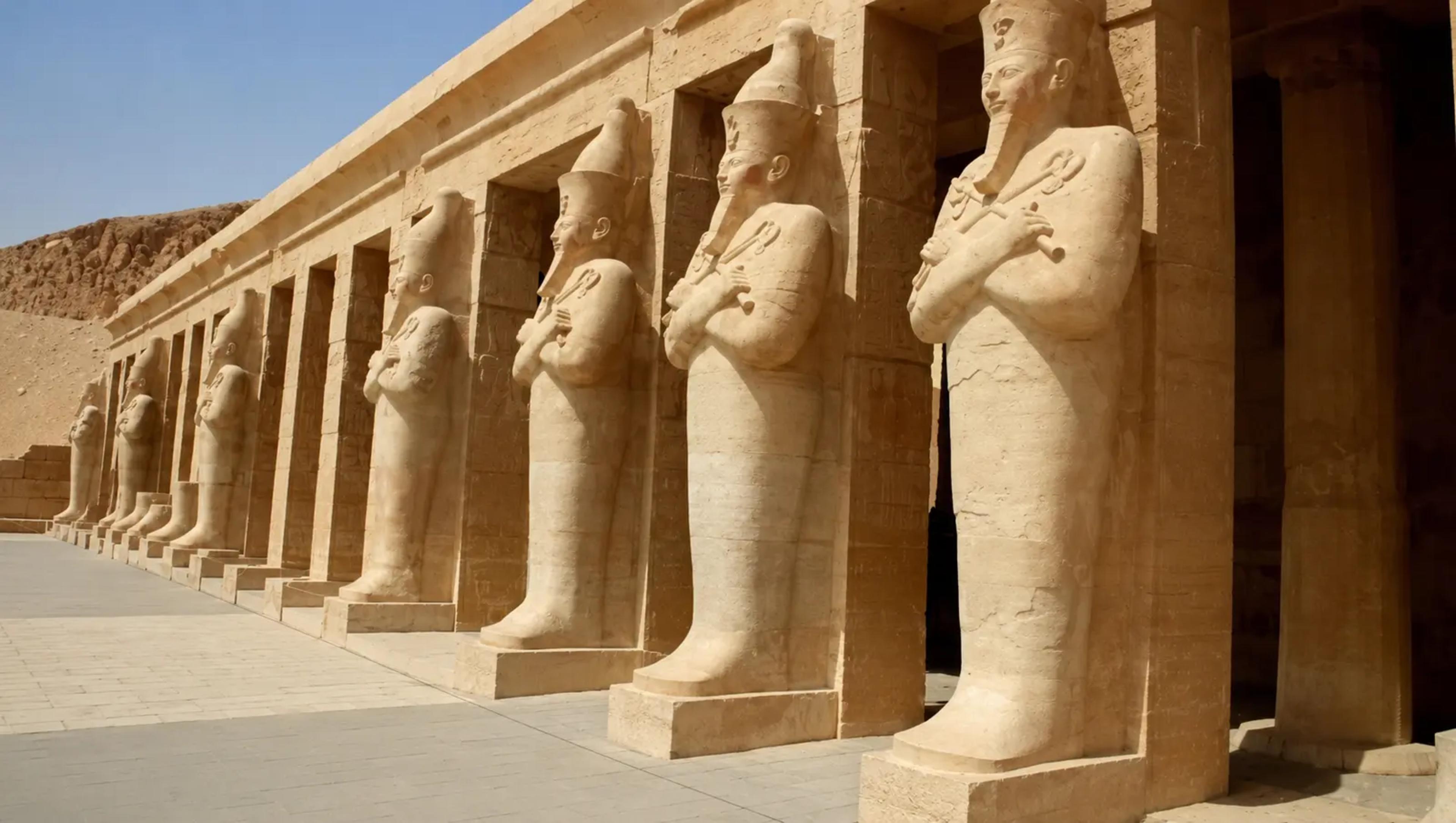 Row of Osiride statues of Queen Hatshepsut along the upper-terrace colonnade of her mortuary temple at Deir el-Bahri, each figure carved against a pillar with arms crossed holding crook and flail.