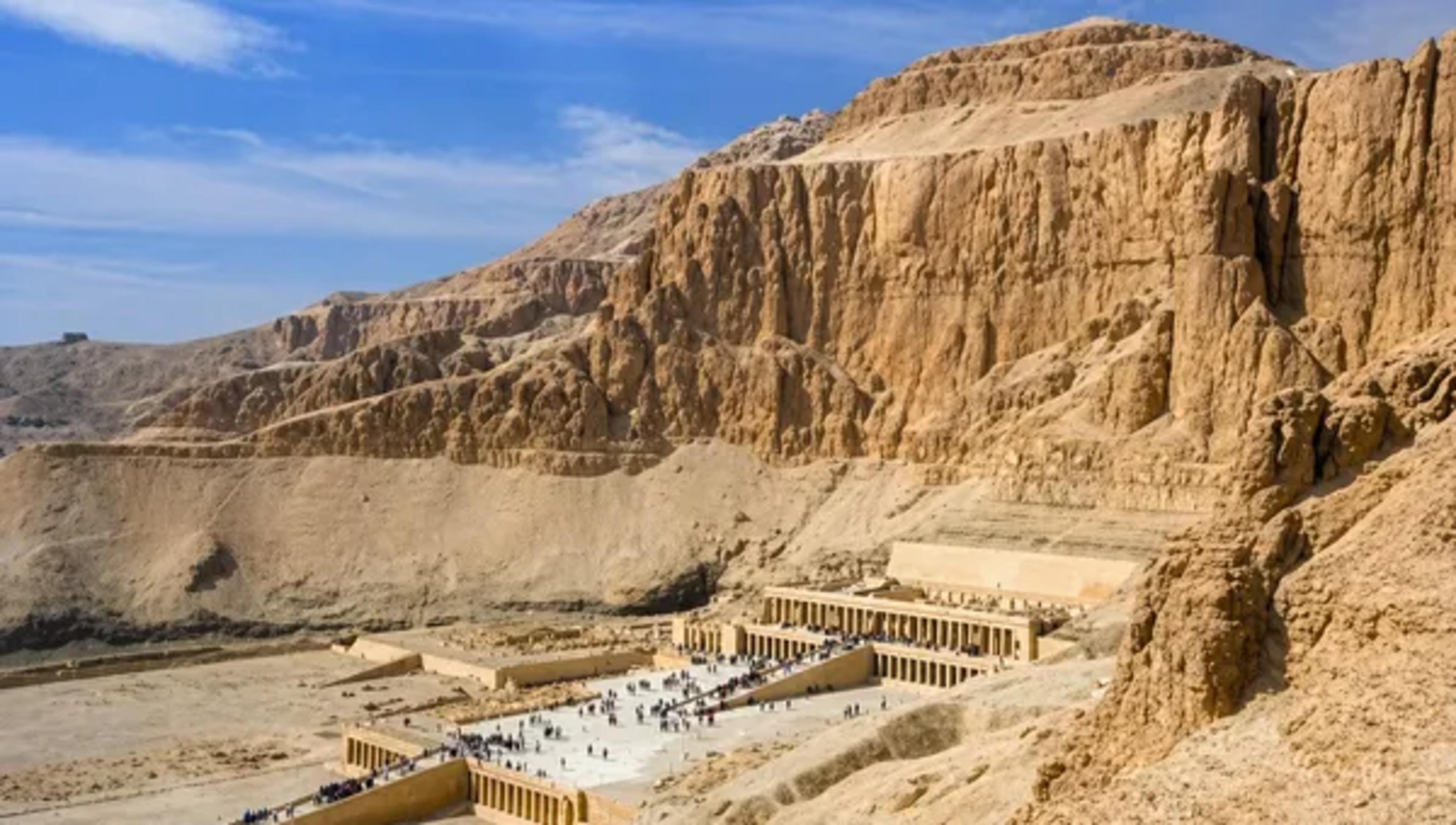 Wide view of the Temple of Hatshepsut at Deir el-Bahri, its three colonnaded terraces and central causeway dwarfed by the sheer limestone cliffs of the Theban necropolis, with crowds of visitors on the approach.