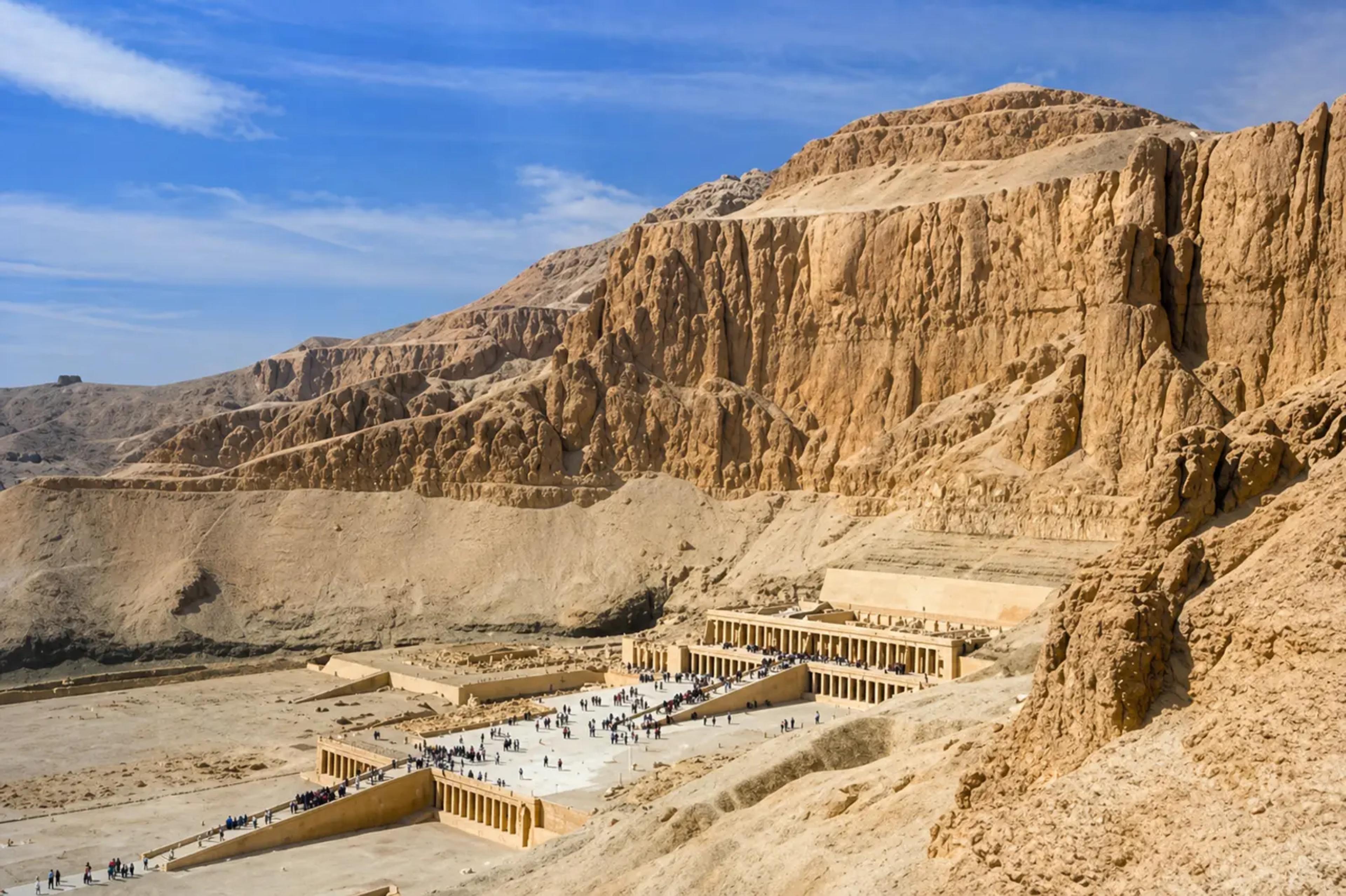Wide view of the Temple of Hatshepsut at Deir el-Bahri, its three colonnaded terraces and central causeway dwarfed by the sheer limestone cliffs of the Theban necropolis, with crowds of visitors on the approach.