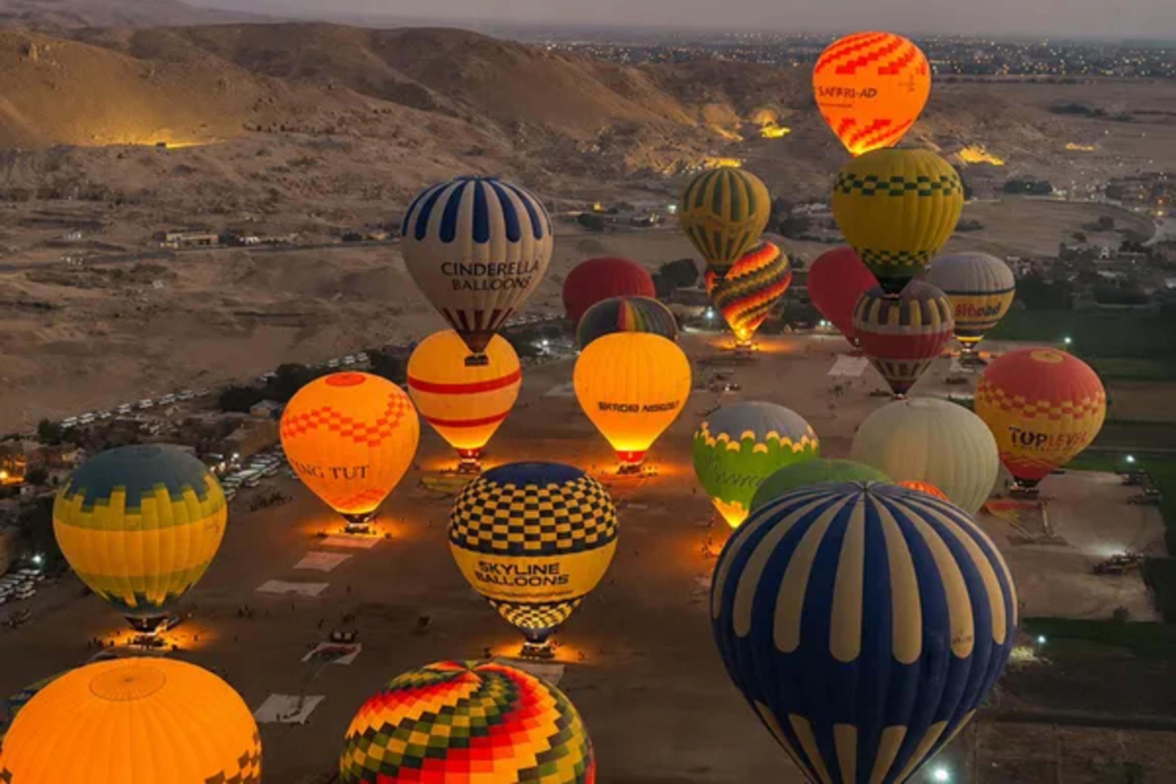 Pre-dawn hot-air balloon launch on the West Bank of Luxor, with dozens of glowing, brightly colored balloons inflating on the desert plain at the foot of the Theban hills.