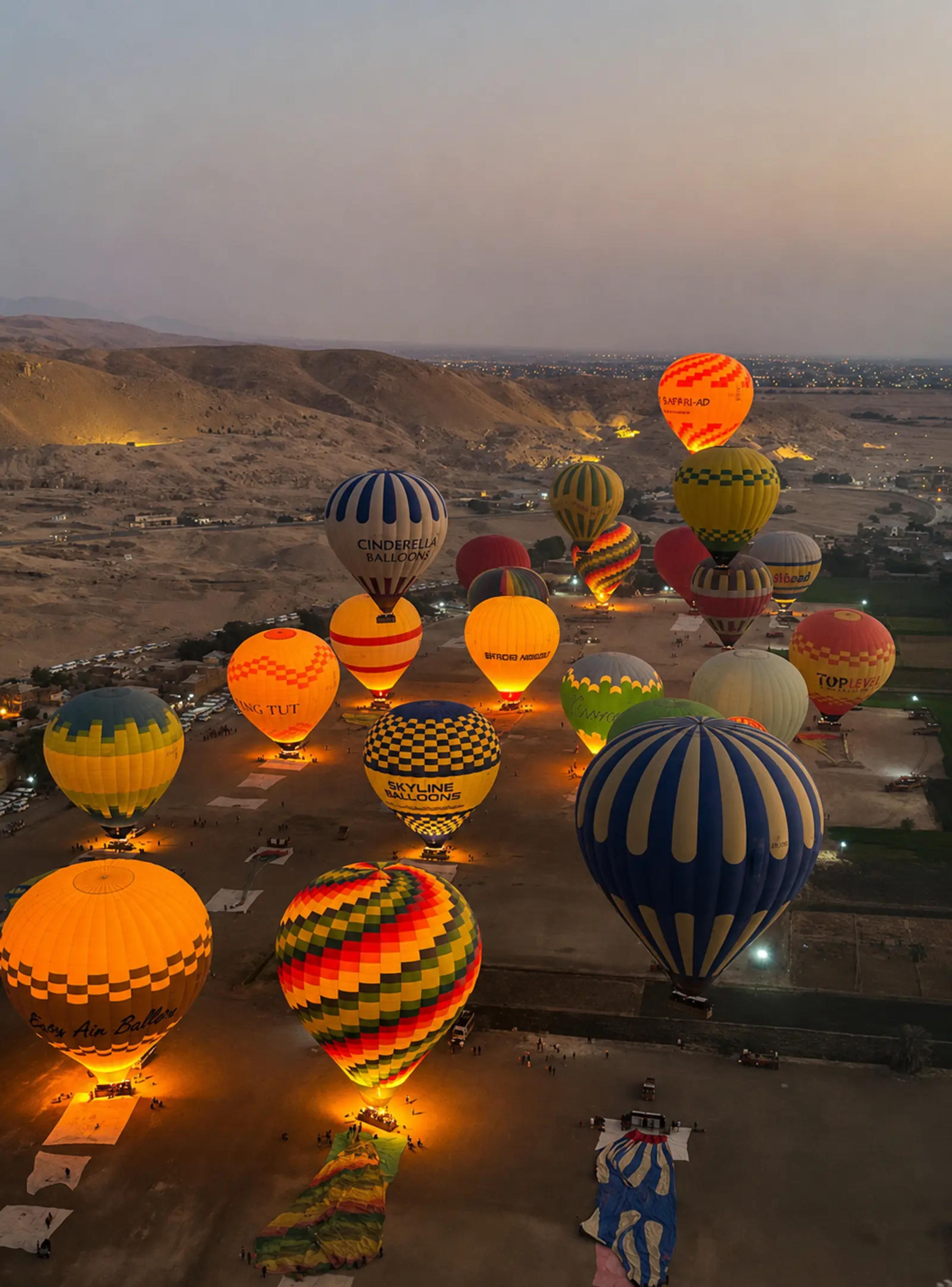 Pre-dawn hot-air balloon launch on the West Bank of Luxor, with dozens of glowing, brightly colored balloons inflating on the desert plain at the foot of the Theban hills.
