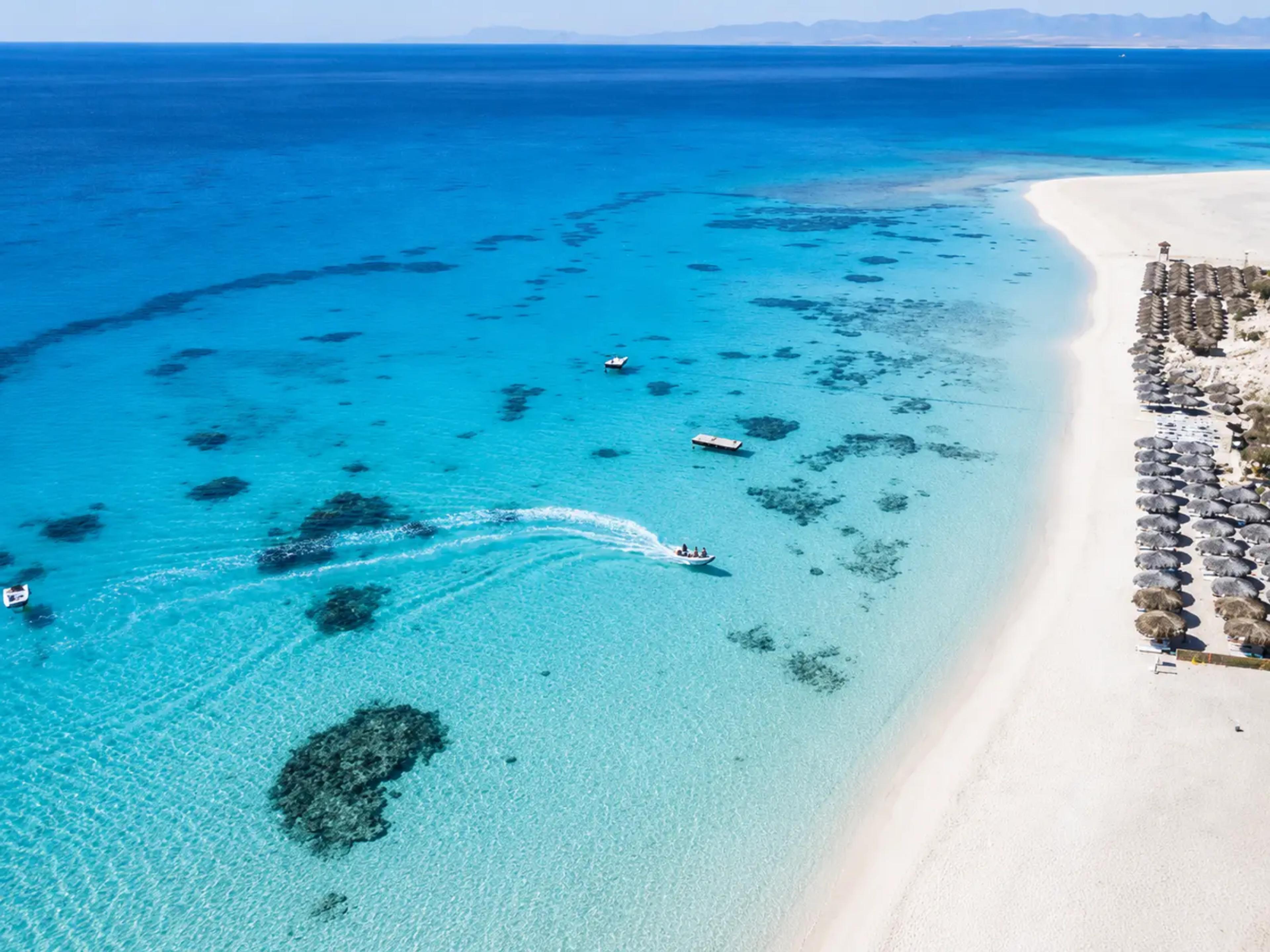 Aerial view of a Hurghada beach on the Red Sea, with rows of straw parasols along white sand, turquoise shallows scattered with dark coral patches, and a small motorboat cutting across the bay.