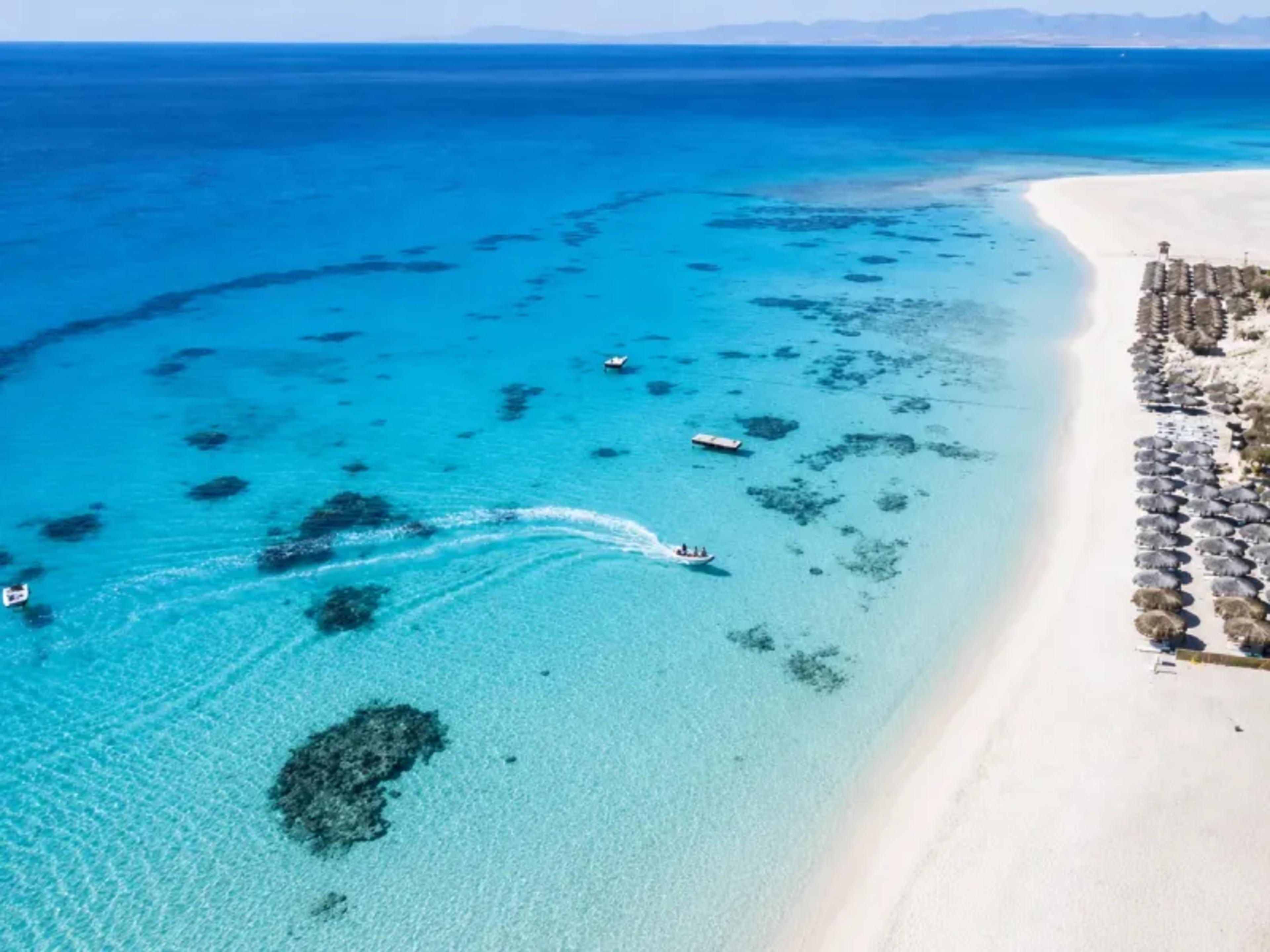 Aerial view of a Hurghada beach on the Red Sea, with rows of straw parasols along white sand, turquoise shallows scattered with dark coral patches, and a small motorboat cutting across the bay.