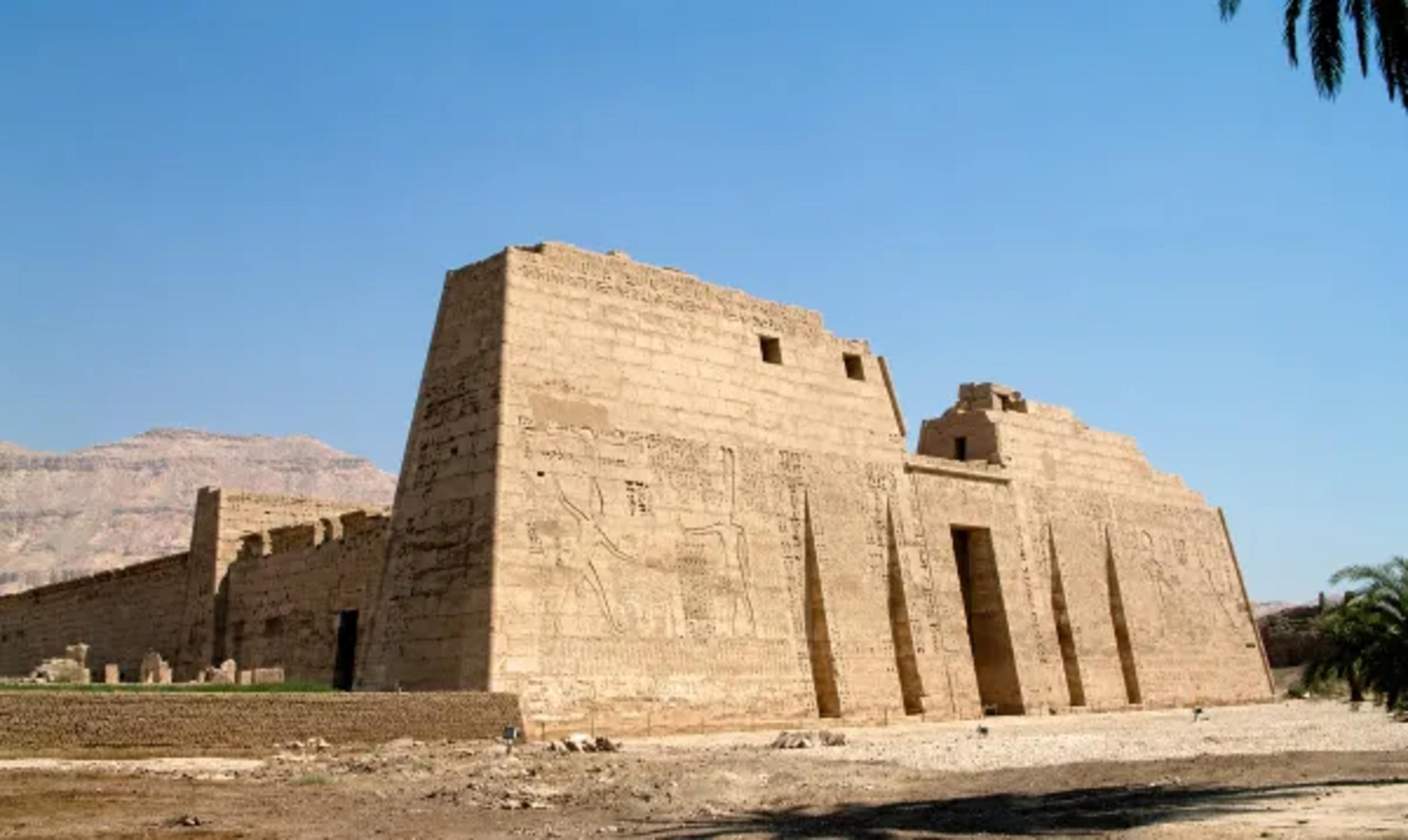 First pylon of Medinet Habu — the mortuary temple of Ramesses III on the West Bank of Luxor — its massive sandstone walls carved with battle reliefs of the pharaoh smiting his enemies, with the Theban hills to the left.
