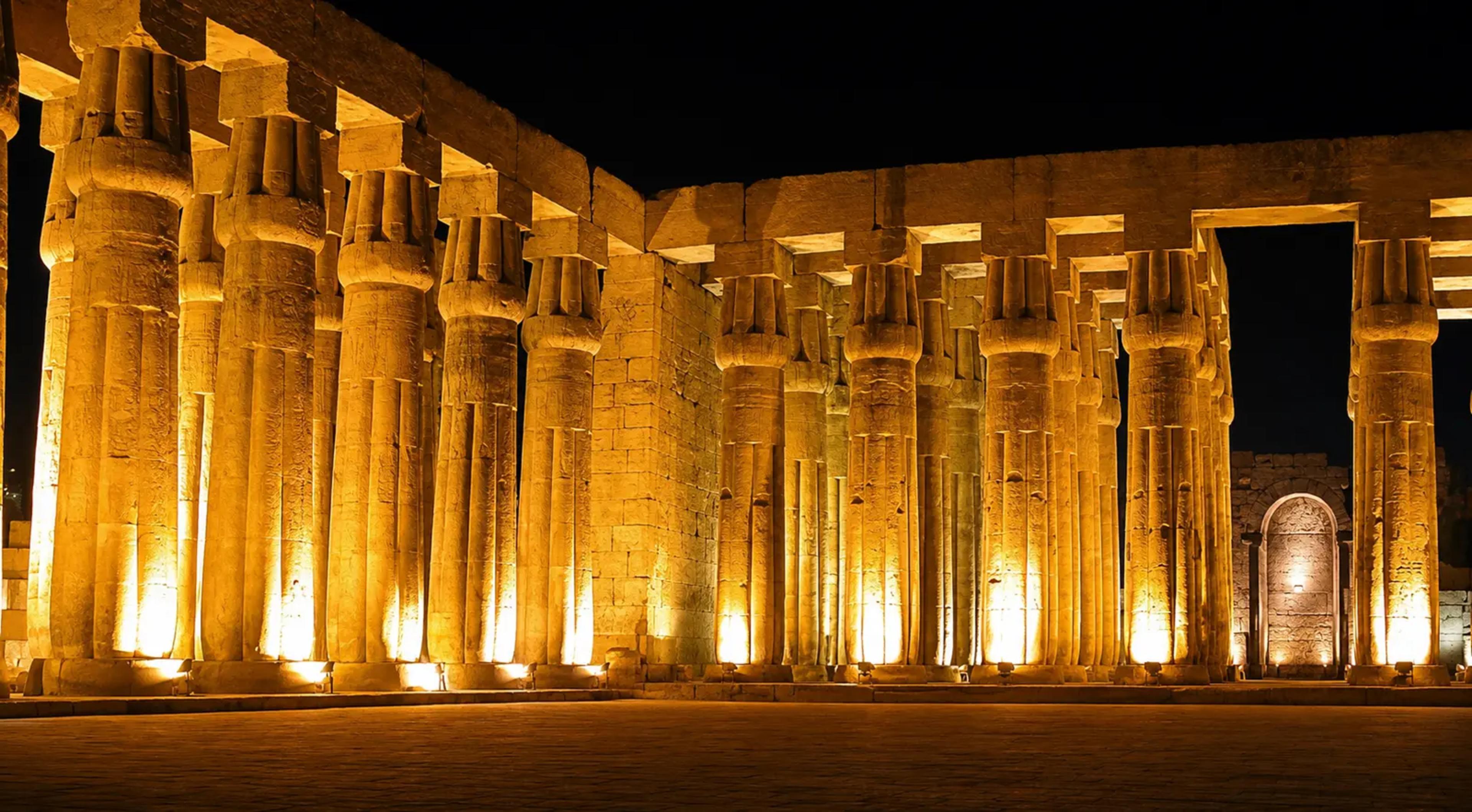 Sun court of Luxor Temple at night, its papyrus-bundle columns uplit in warm gold against a black sky, with a Roman-era apsidal niche visible at the rear wall.