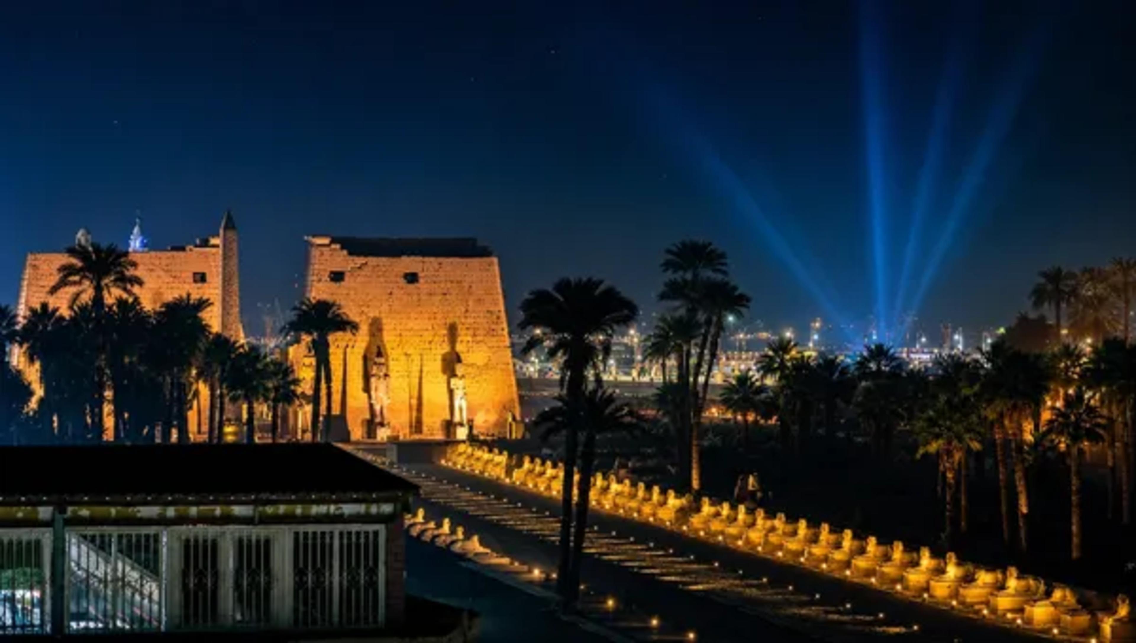 Illuminated ancient Egyptian temple and palm-lined avenue at night with blue light beams in the sky.