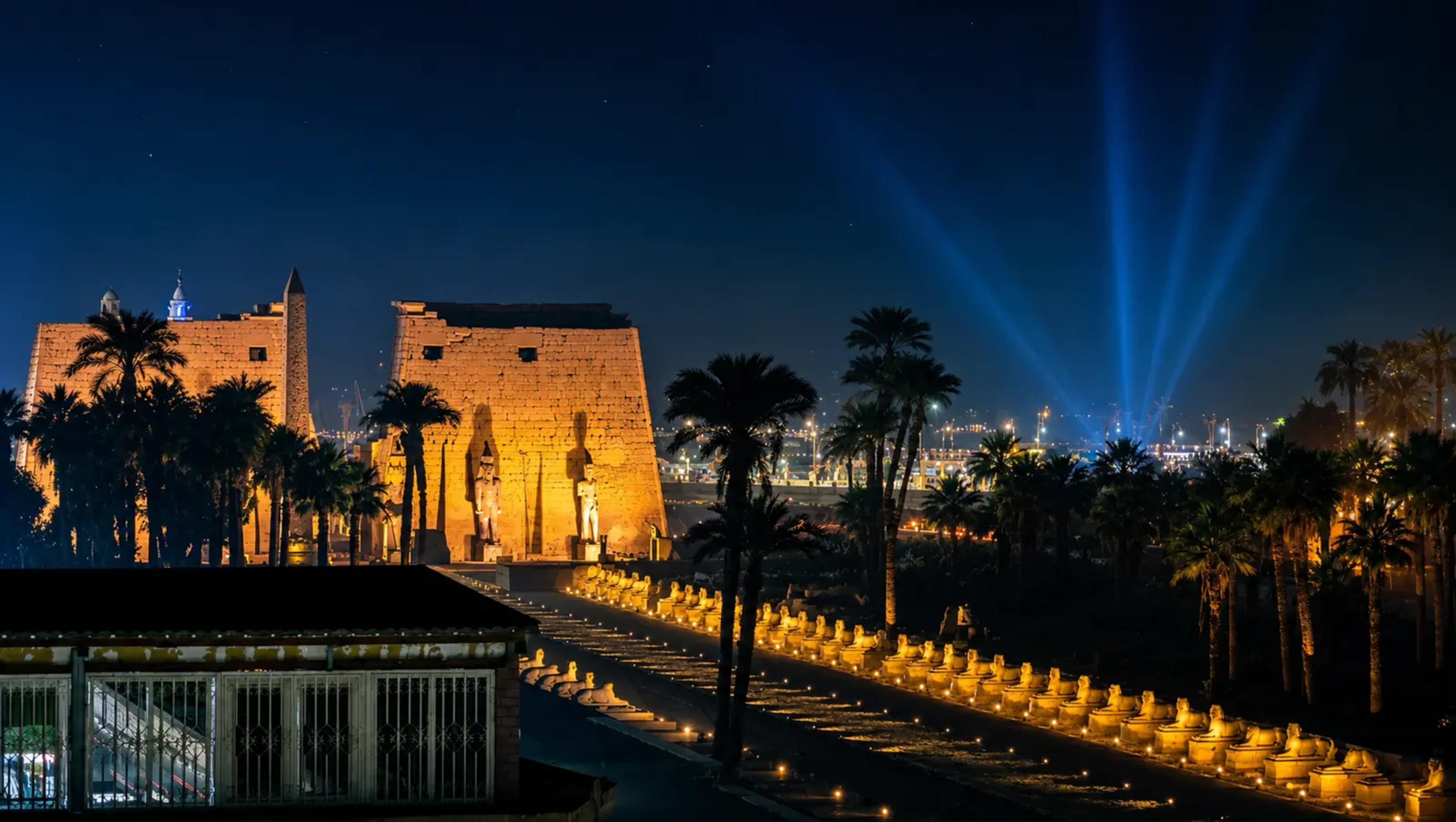 Illuminated ancient Egyptian temple and palm-lined avenue at night with blue light beams in the sky.