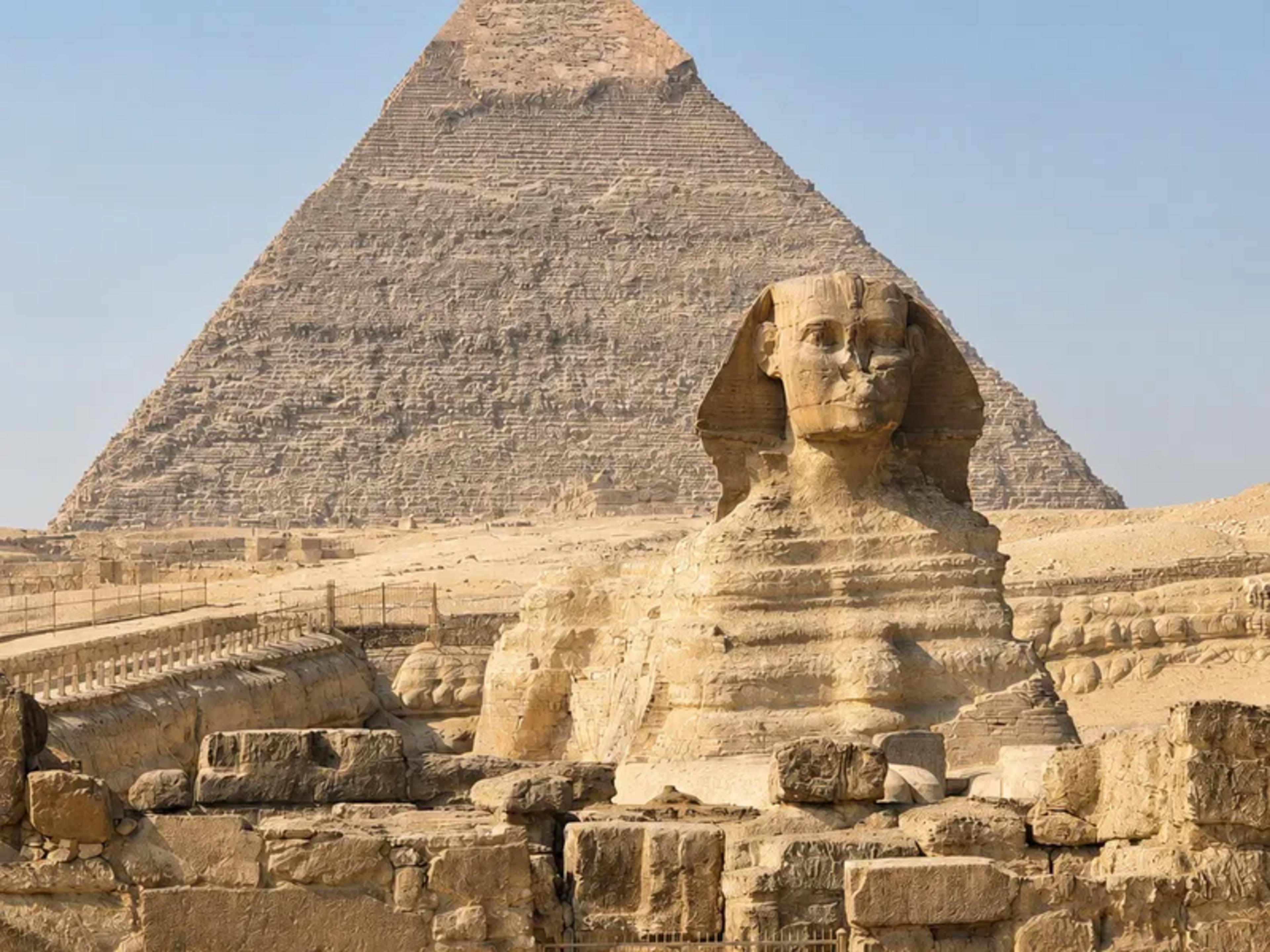 The Great Sphinx of Giza in the foreground, its weathered limestone body and pharaoh's head facing the camera, with the Pyramid of Khafre rising directly behind and small groups of visitors at the base for scale.