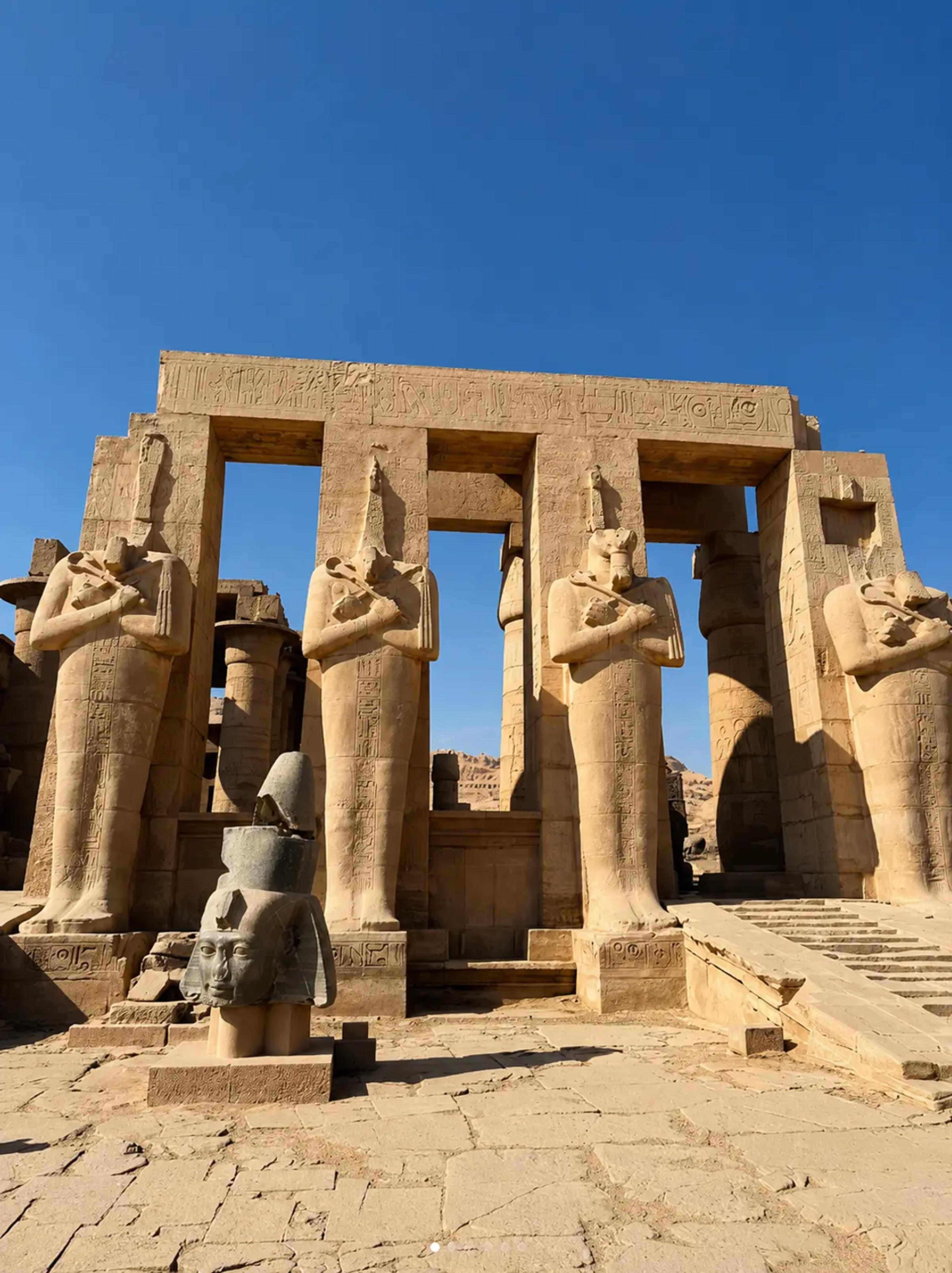 Second courtyard of the Ramesseum on the West Bank of Luxor, with a row of Osiride pillars depicting Ramesses II as Osiris flanking the entrance and a fallen granite head of the king in the foreground against a clear blue sky.