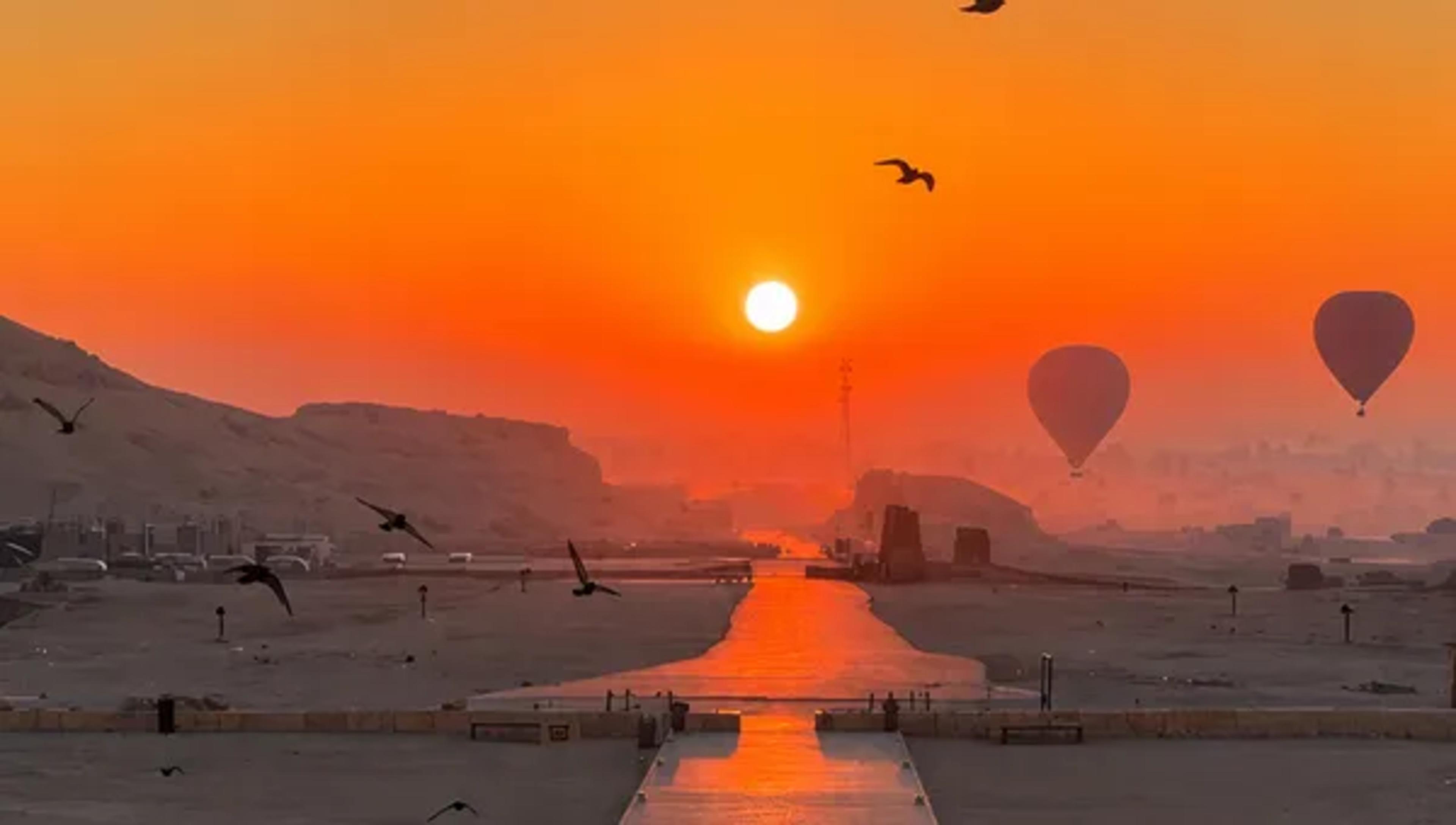 Sunrise over the Avenue of Sphinxes between Karnak and Luxor Temples, with the sun rising at the end of the stone causeway, silhouetted hot-air balloons drifting on the right, and birds in flight against a deep-orange sky.
