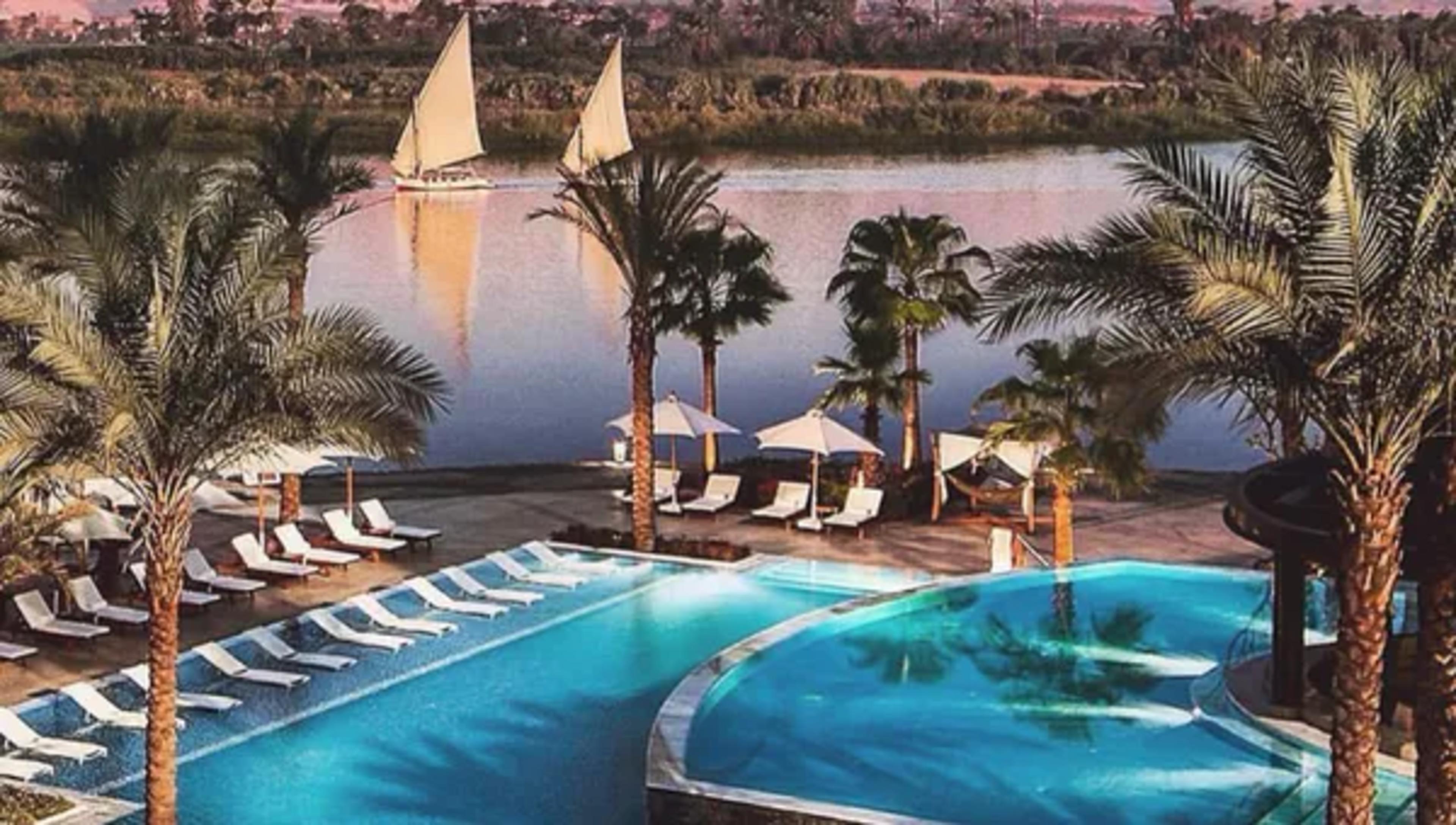 Luxor riverside hotel pool fringed with date palms and sun loungers, looking across the Nile to a felucca under sail and the pink-lit Theban hills at sunset.