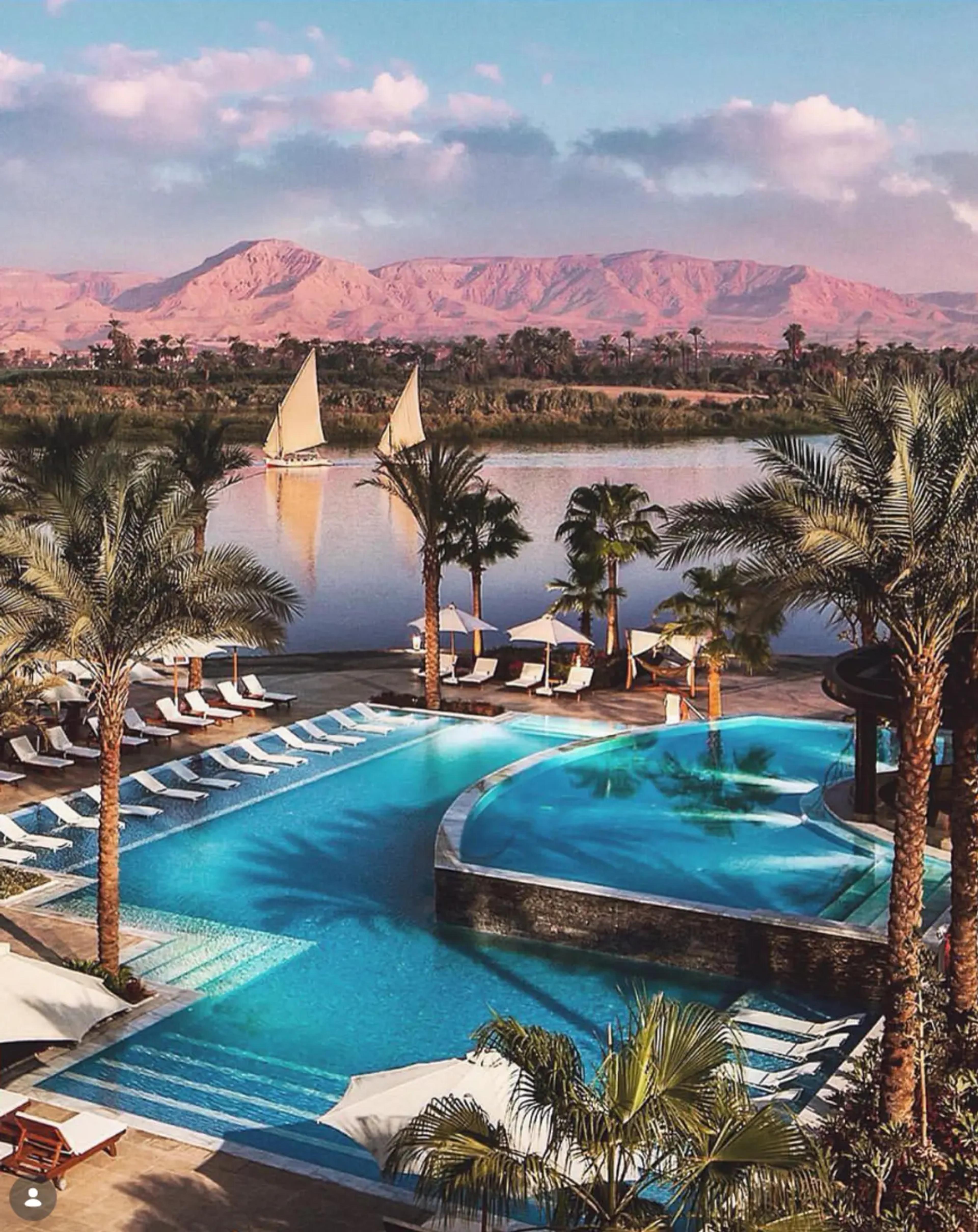 Luxor riverside hotel pool fringed with date palms and sun loungers, looking across the Nile to a felucca under sail and the pink-lit Theban hills at sunset.