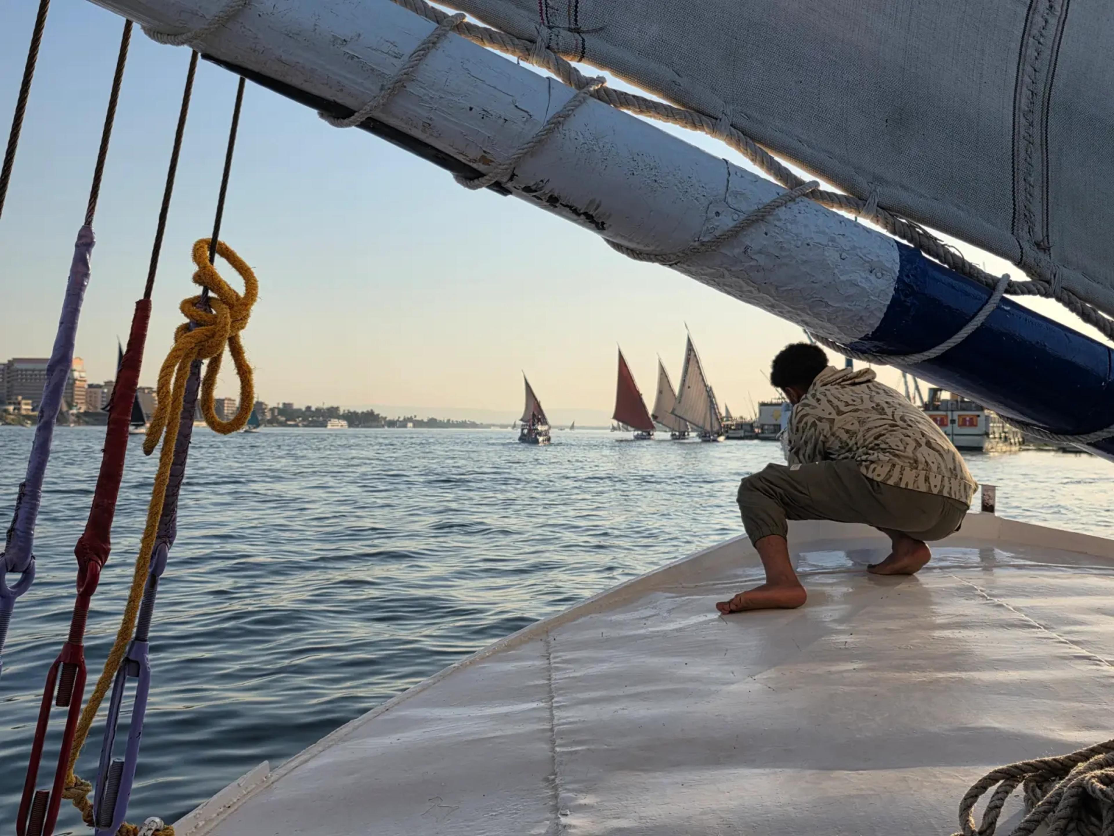 A felucca crewman raising the sail on the Nile at sunset, with a fleet of feluccas drifting downriver toward Luxor's east bank
