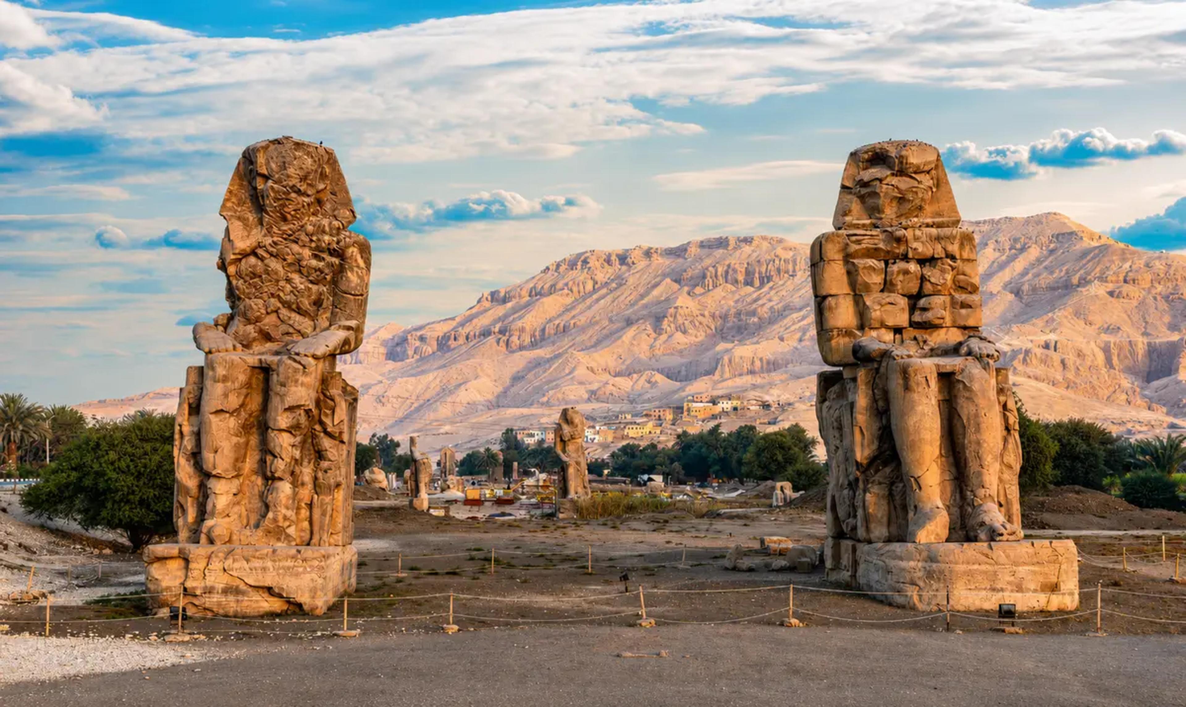 The two seated Colossi of Memnon — weathered sandstone statues of Pharaoh Amenhotep III — flanking the entrance to his mortuary temple on the West Bank of Luxor, with the limestone Theban hills behind.