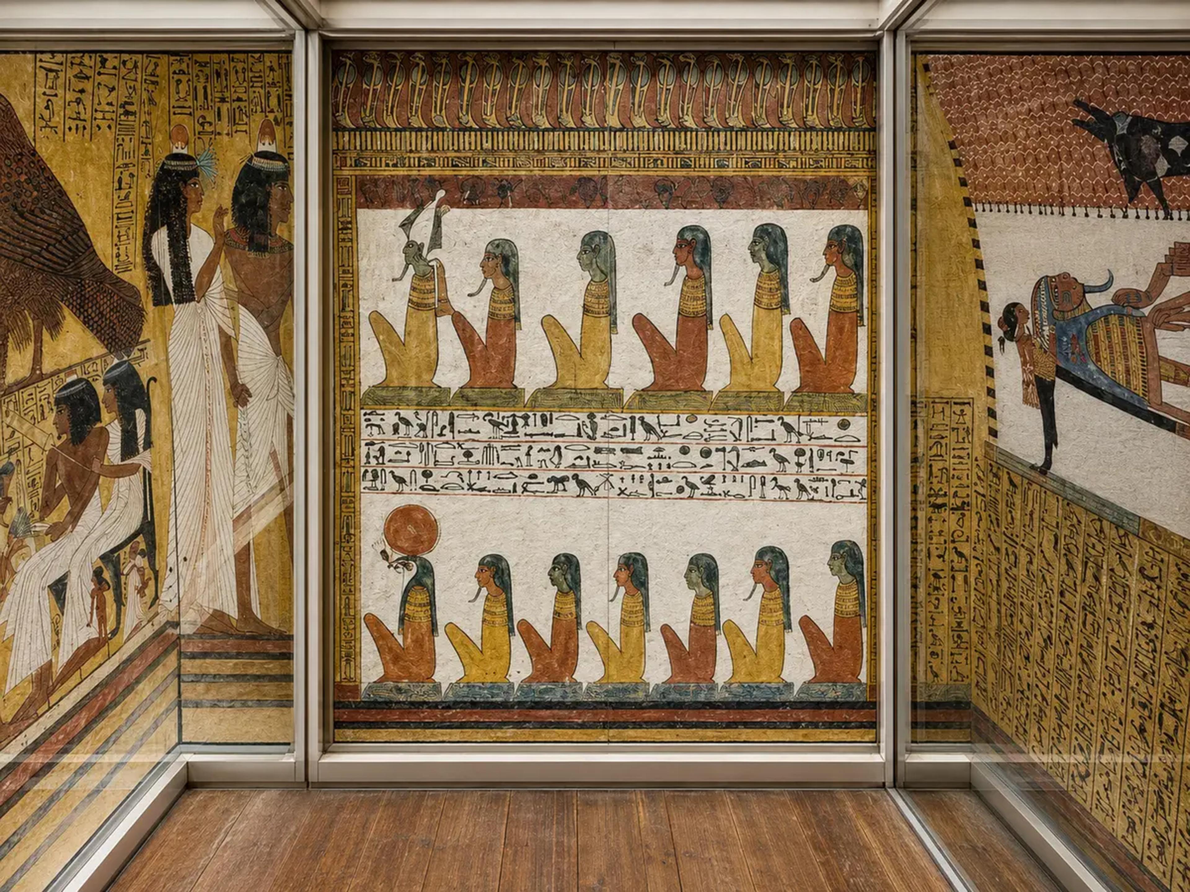 Straight-on view of painted wall scenes and hieroglyphs from the Valley of the Nobles, displayed behind glass with a wooden floor in the foreground.