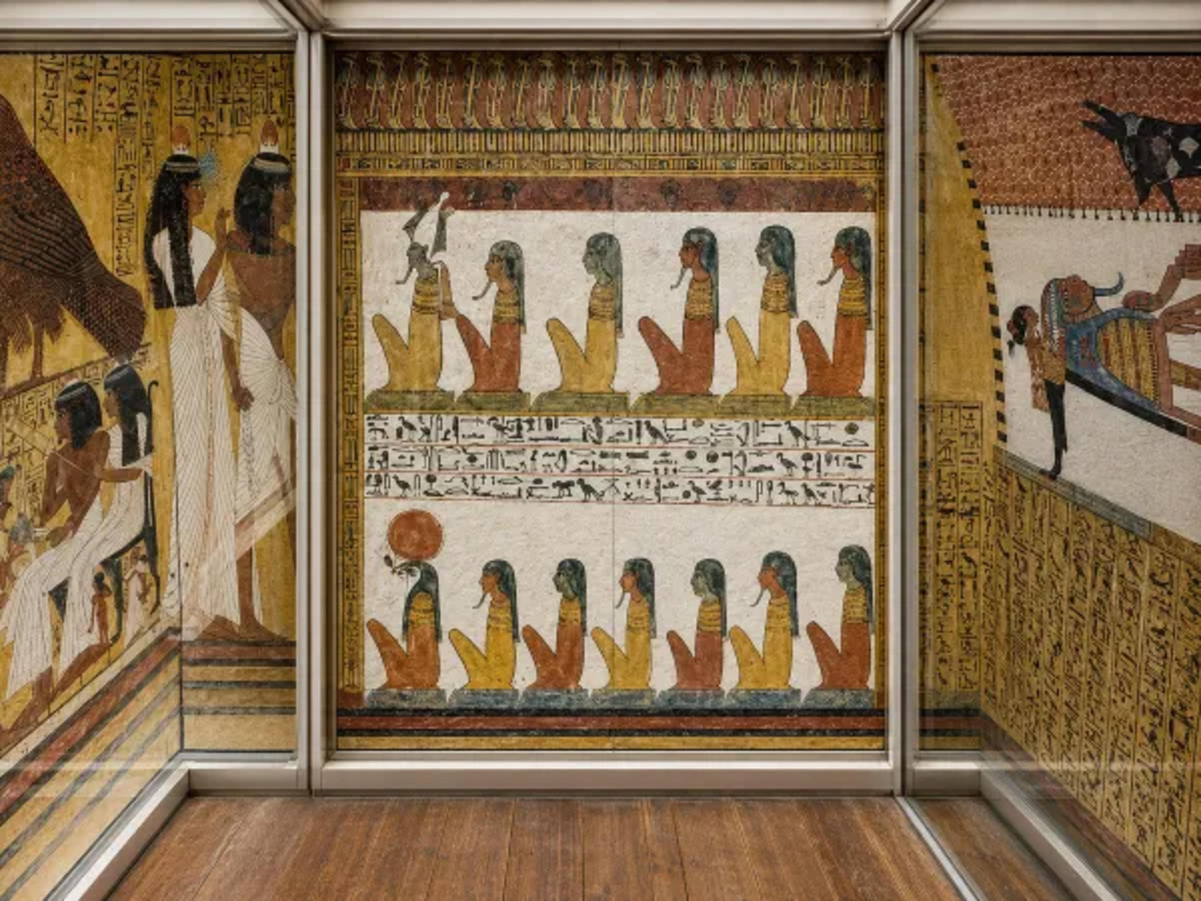 Straight-on view of painted wall scenes and hieroglyphs from the Valley of the Nobles, displayed behind glass with a wooden floor in the foreground.