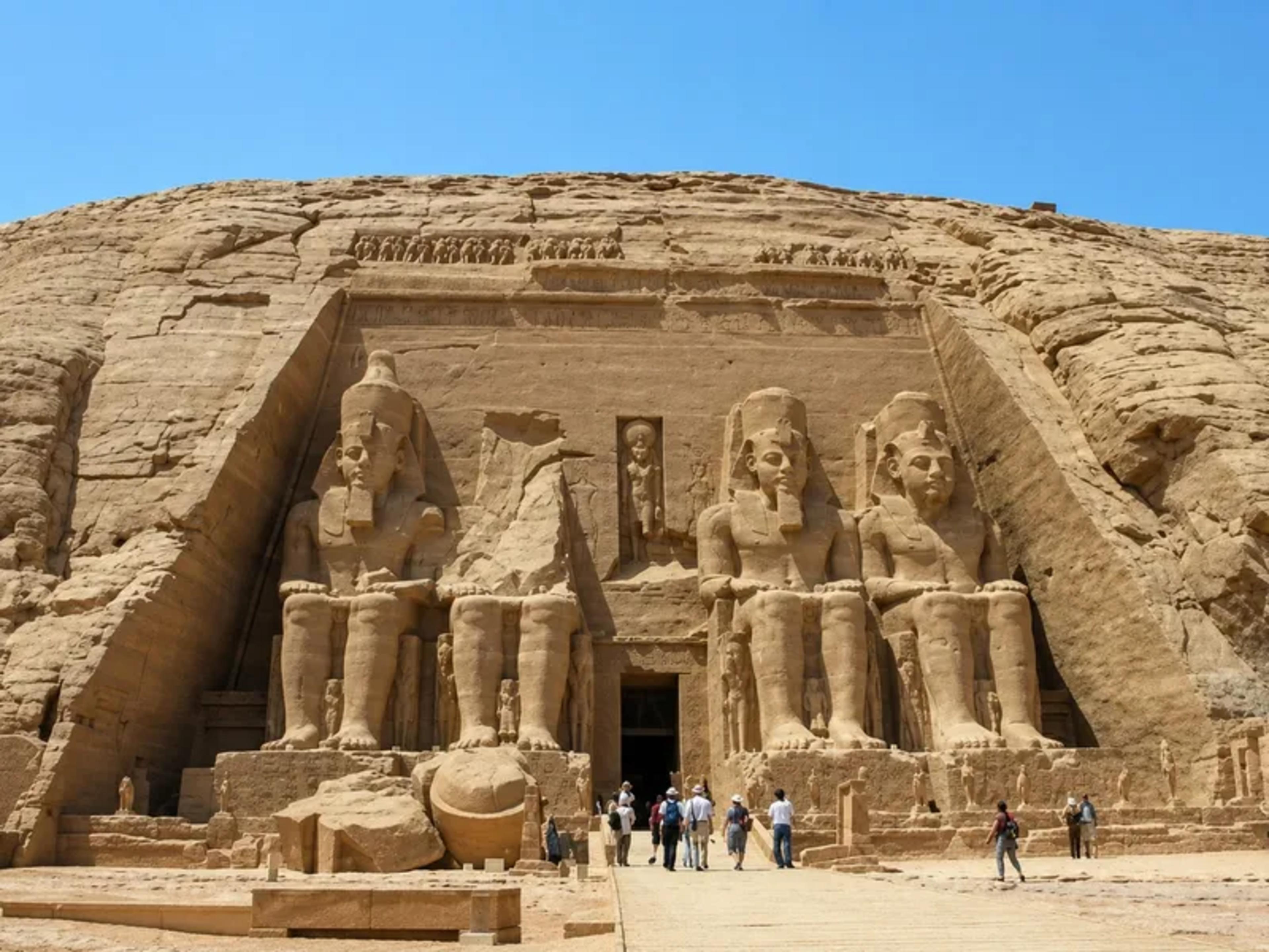 Façade of the Great Temple of Abu Simbel, with four colossal seated statues of Ramesses II carved into the sandstone cliff and visitors approaching the central entrance for scale, under a clear blue sky.