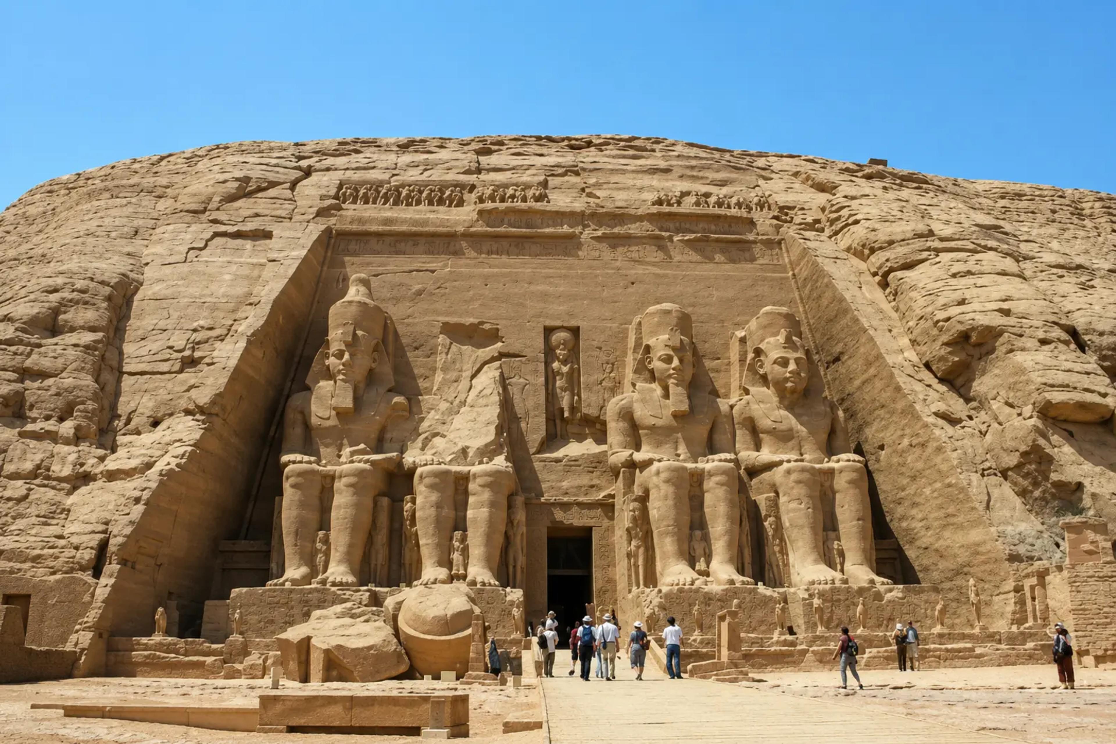 Façade of the Great Temple of Abu Simbel, with four colossal seated statues of Ramesses II carved into the sandstone cliff and visitors approaching the central entrance for scale, under a clear blue sky.