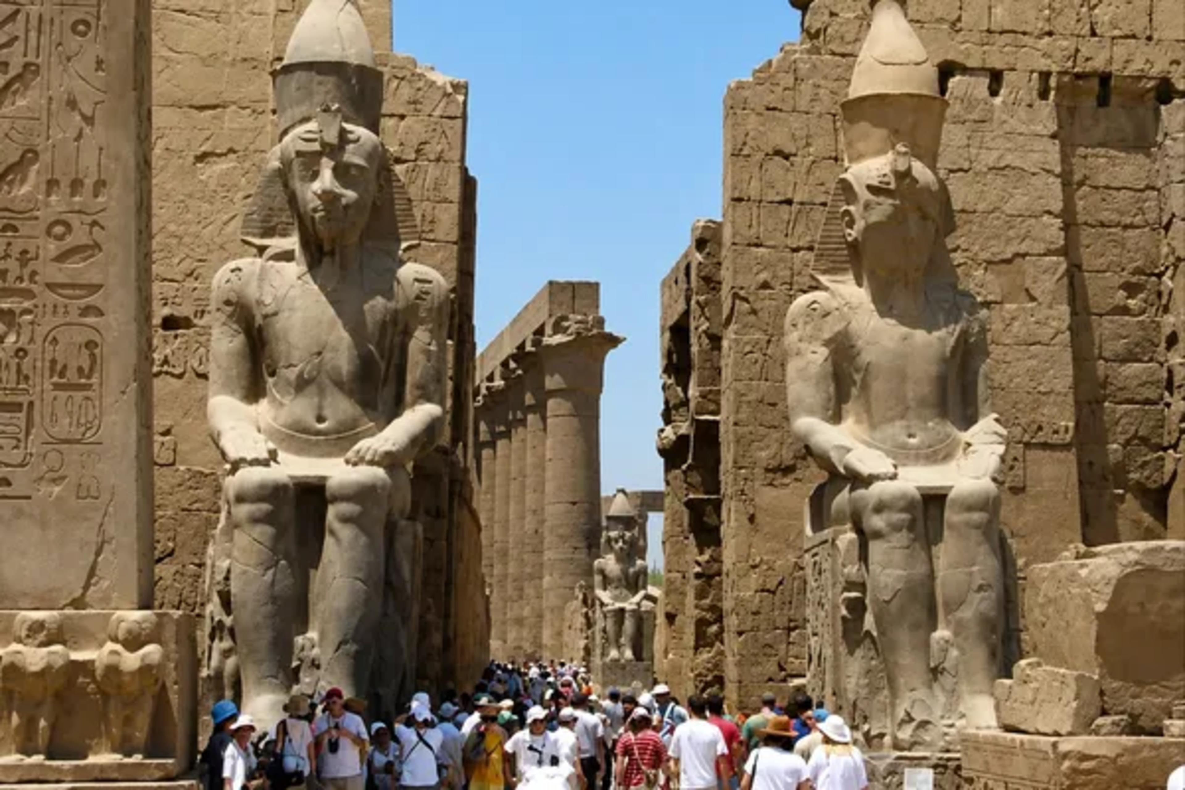 Crowds passing between the colossal seated statues of Ramesses II at the entrance pylon of Luxor Temple, with the obelisk and inner colonnade beyond