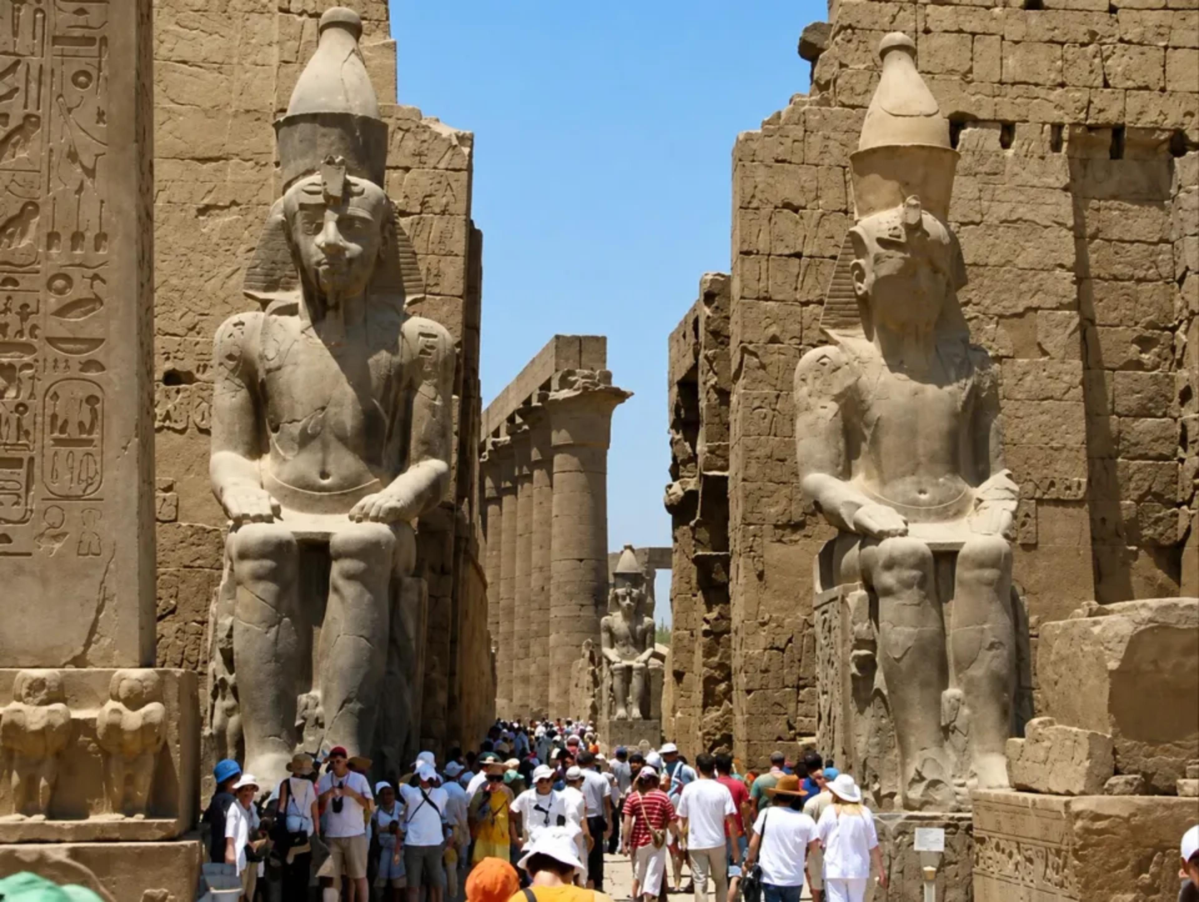 Crowds passing between the colossal seated statues of Ramesses II at the entrance pylon of Luxor Temple, with the obelisk and inner colonnade beyond