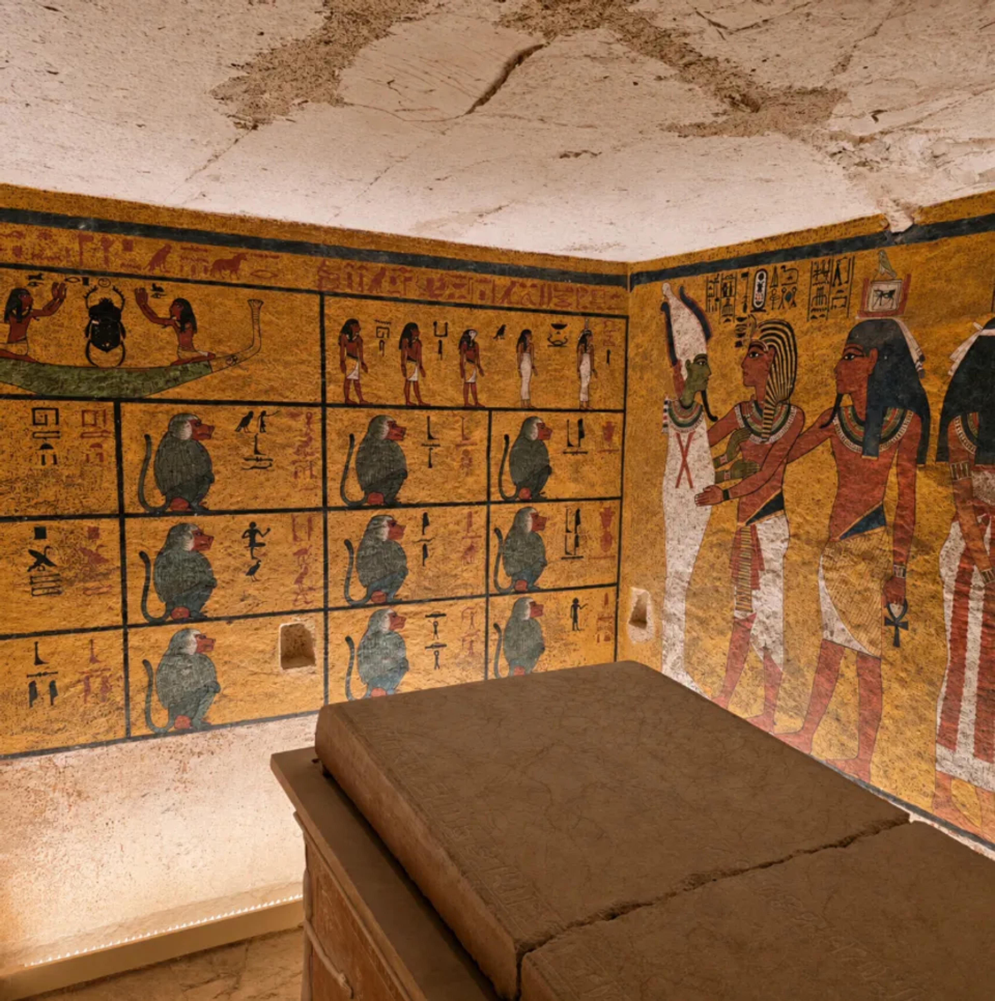Interior of the tomb of Tutankhamun in the Valley of the Kings, with yellow-ground burial-chamber walls showing the twelve baboons of the Amduat and Tutankhamun greeted by Osiris, the king's stone sarcophagus in the foreground.