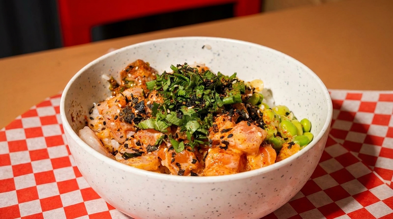 A colorful poke bowl with salmon, edamame, and herbs on a red and white checkered paper.