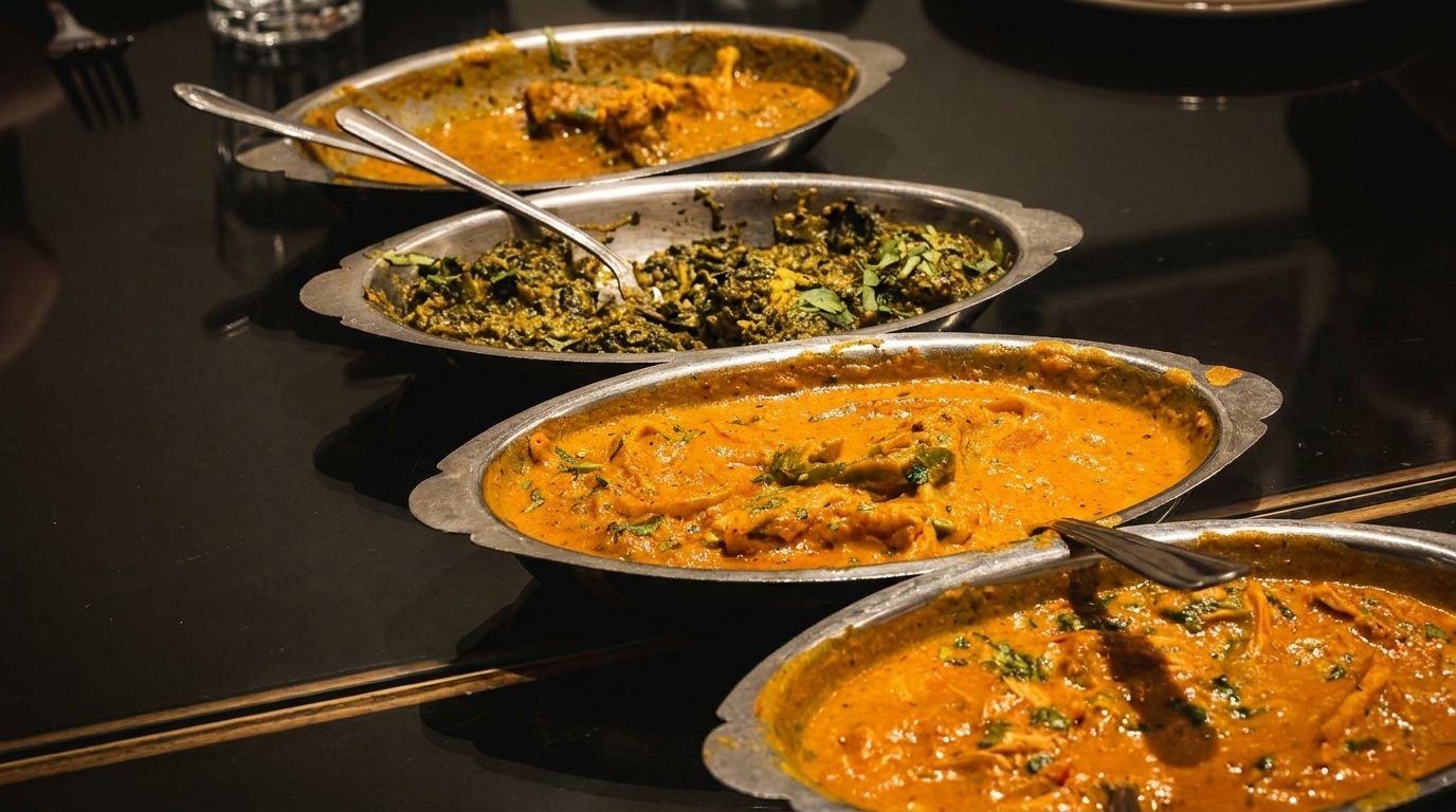 Four oval metal dishes filled with various Indian curries and green vegetable dishes on a dark table.