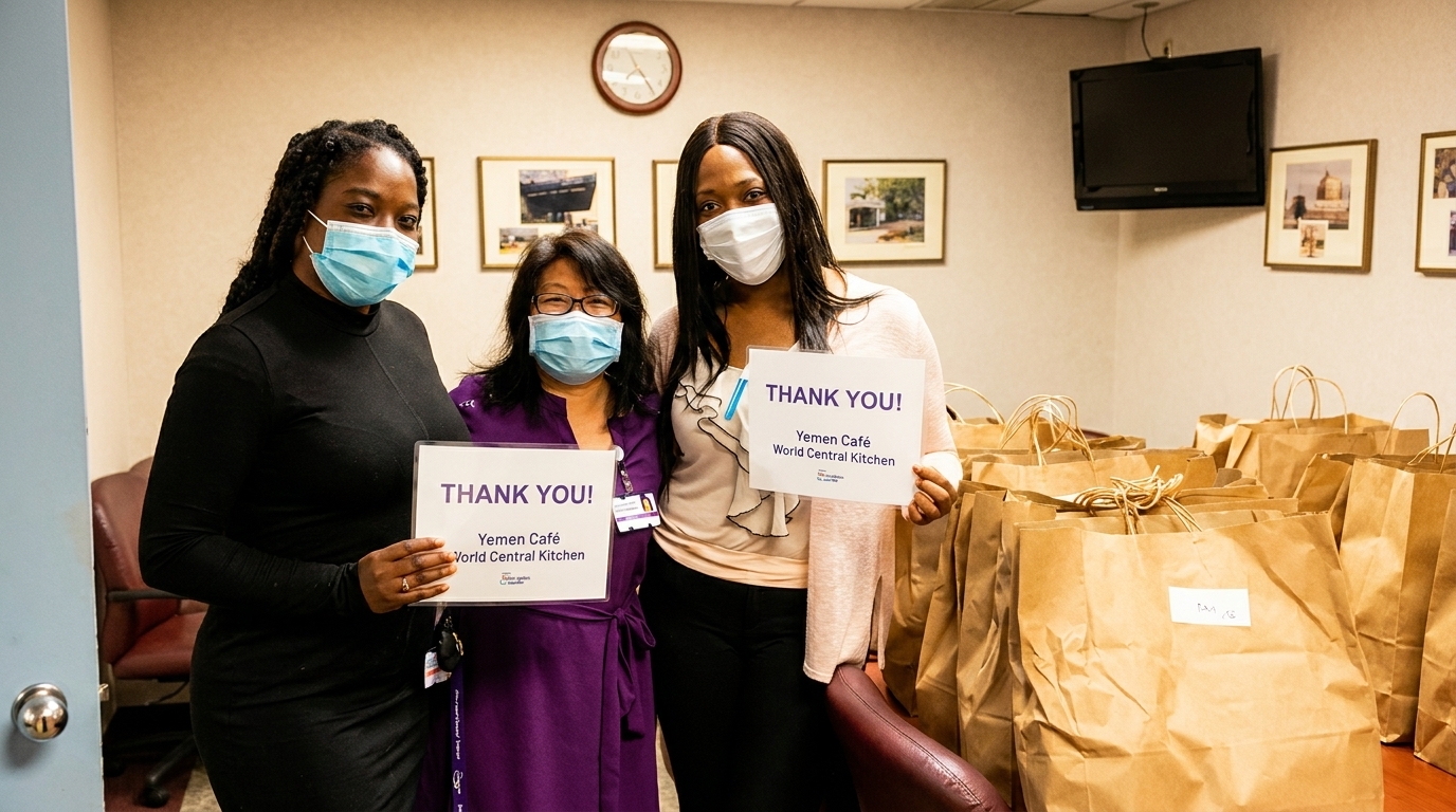 Three women in masks hold "Thank You" signs from Yemen Café and World Central Kitchen, surrounded by many brown paper bags.