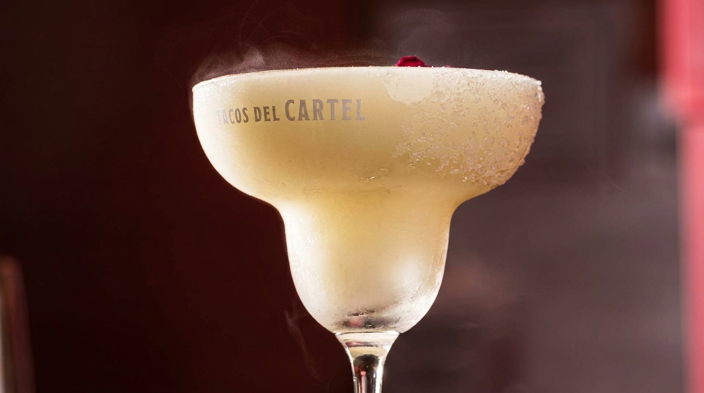 A frosty margarita in a branded glass with a salted rim and a red garnish, emitting vapor.