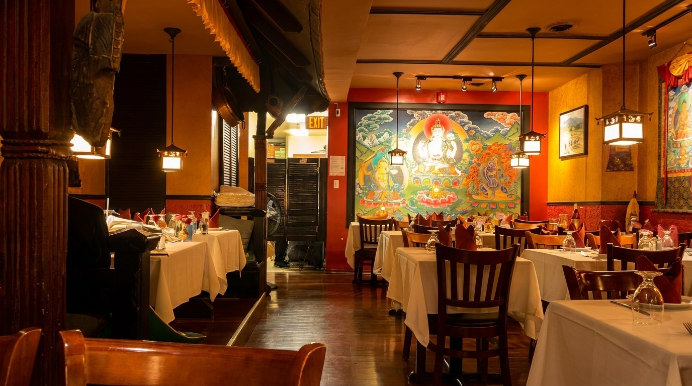 Warmly lit restaurant interior with white-clothed tables, wooden chairs, and colorful Buddhist thangka paintings on red walls.