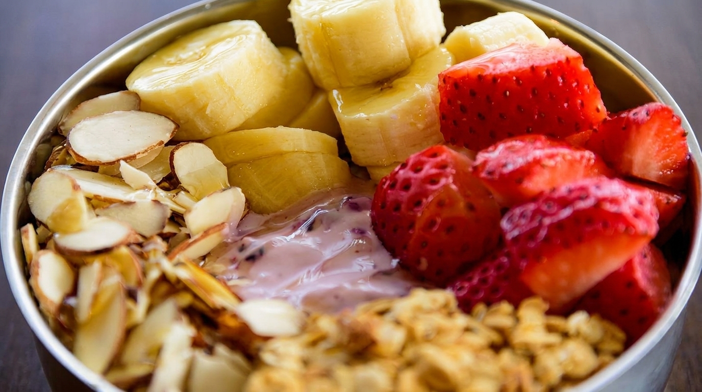Close-up of a breakfast bowl with sliced bananas, strawberries, almonds, granola, and yogurt.