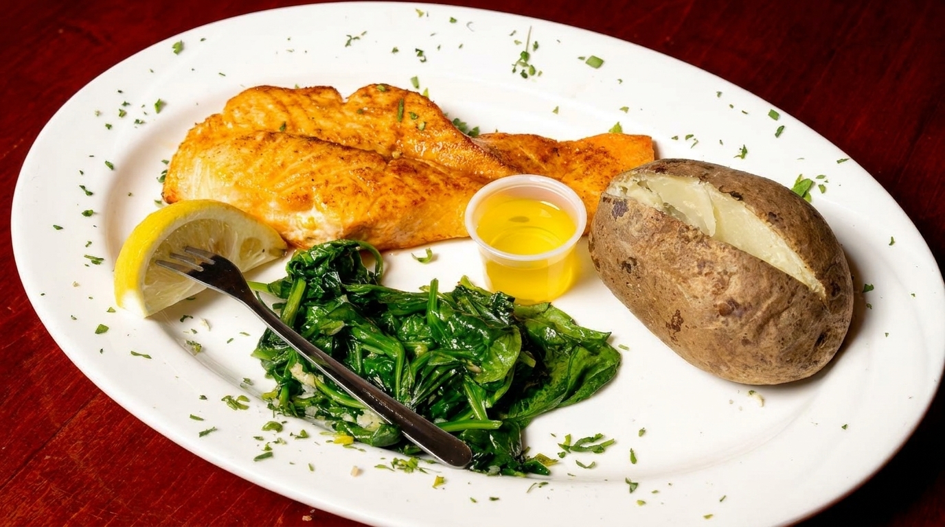 Grilled salmon with a lemon wedge, sautéed spinach, a baked potato, and melted butter on a white plate.