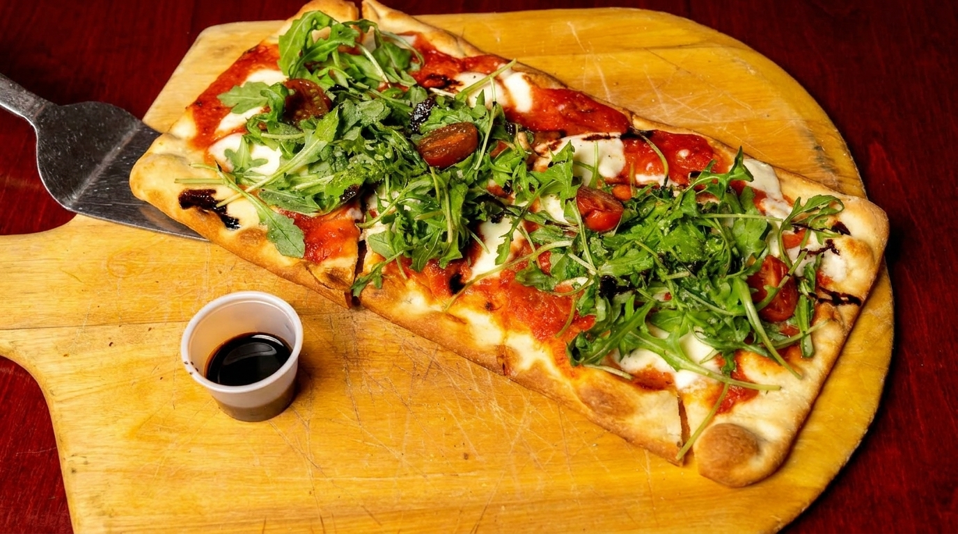 A rectangular flatbread pizza with tomato sauce, mozzarella, arugula, and cherry tomatoes on a wooden peel.