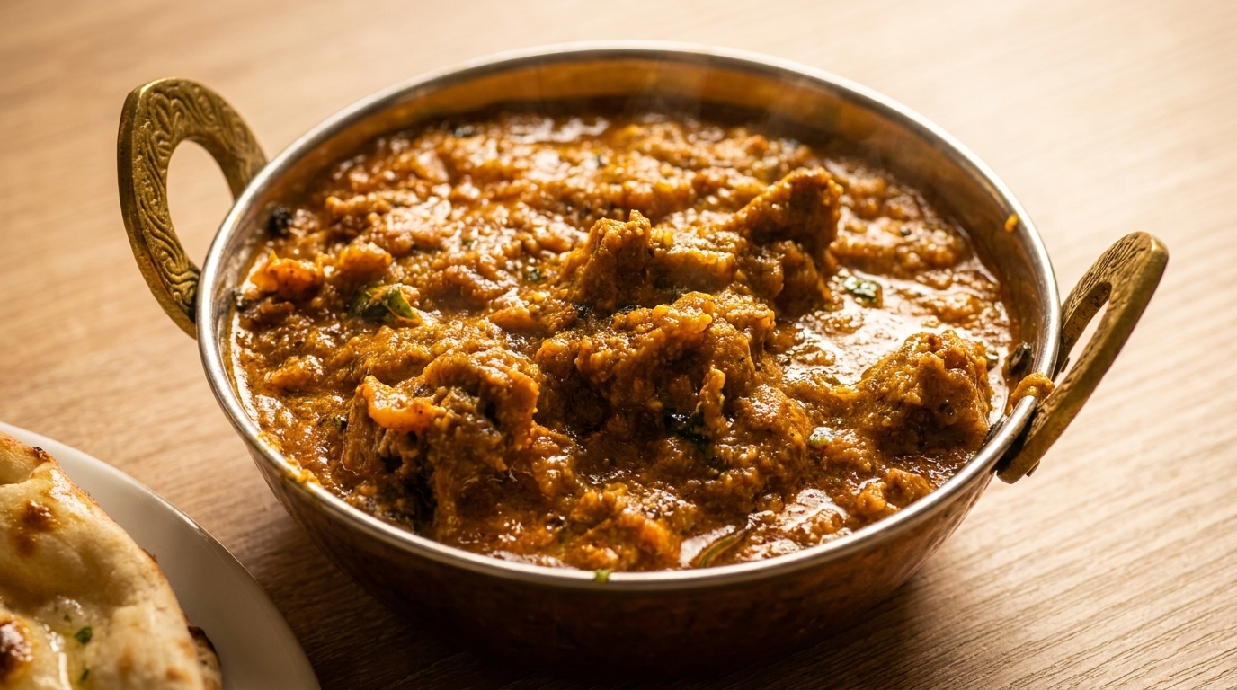 A copper karahi bowl filled with a rich, orange-brown curry and a piece of naan bread.