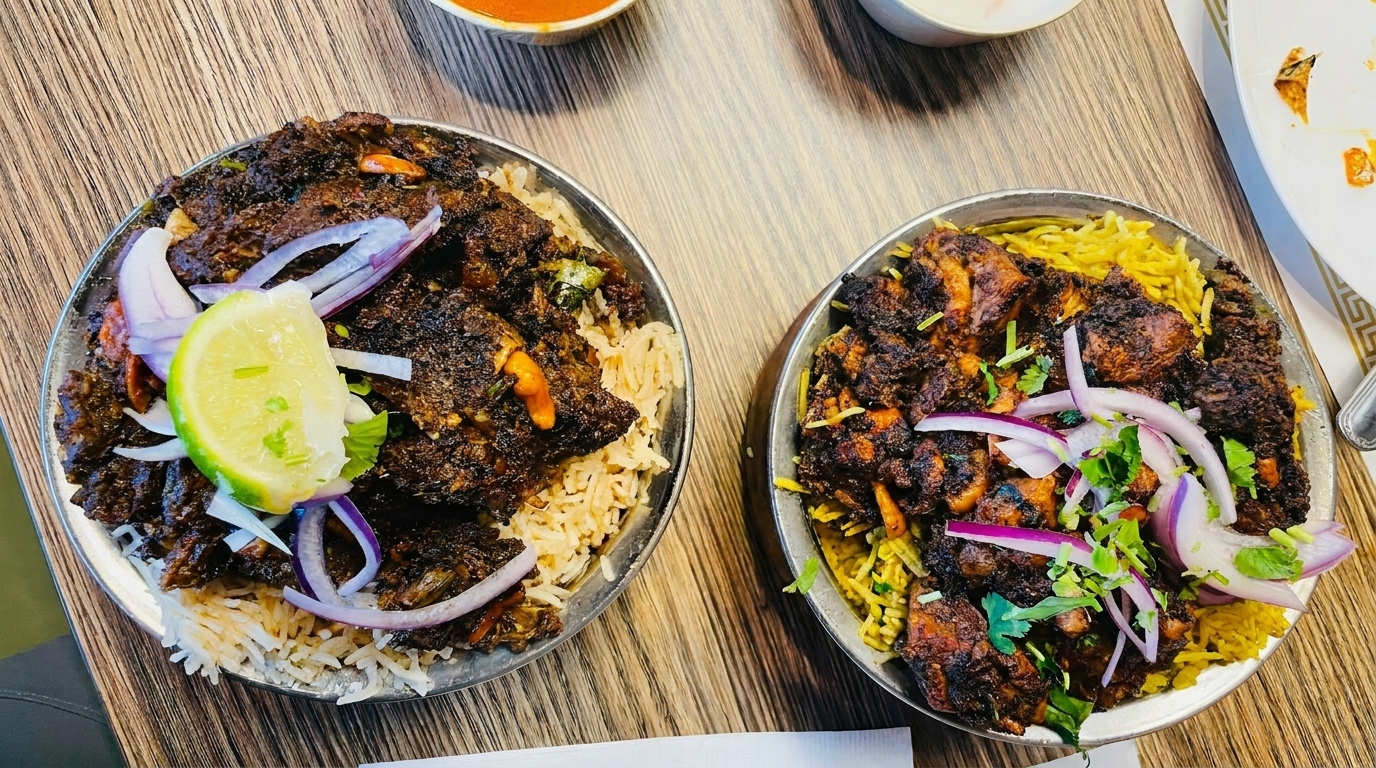 Two metal bowls of dark, spicy chicken biryani with rice, onions, and lime on a wooden table.
