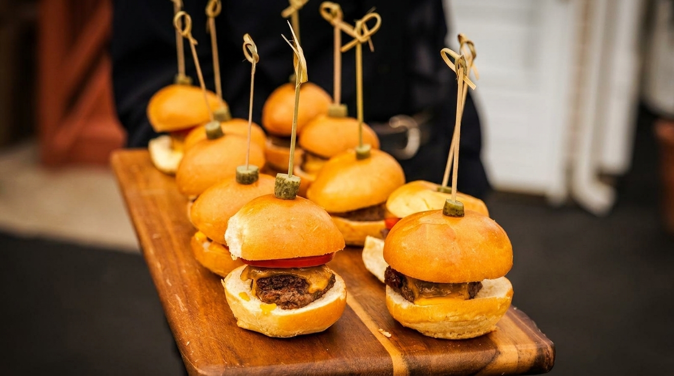 Miniature cheeseburger sliders with pickles on skewers, served on a wooden board.