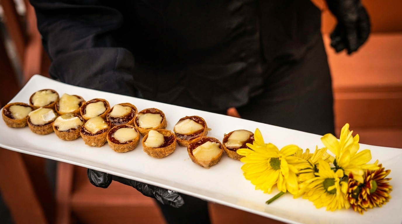 A server in black gloves holds a white tray of mini tarts with melted cheese and yellow flowers.