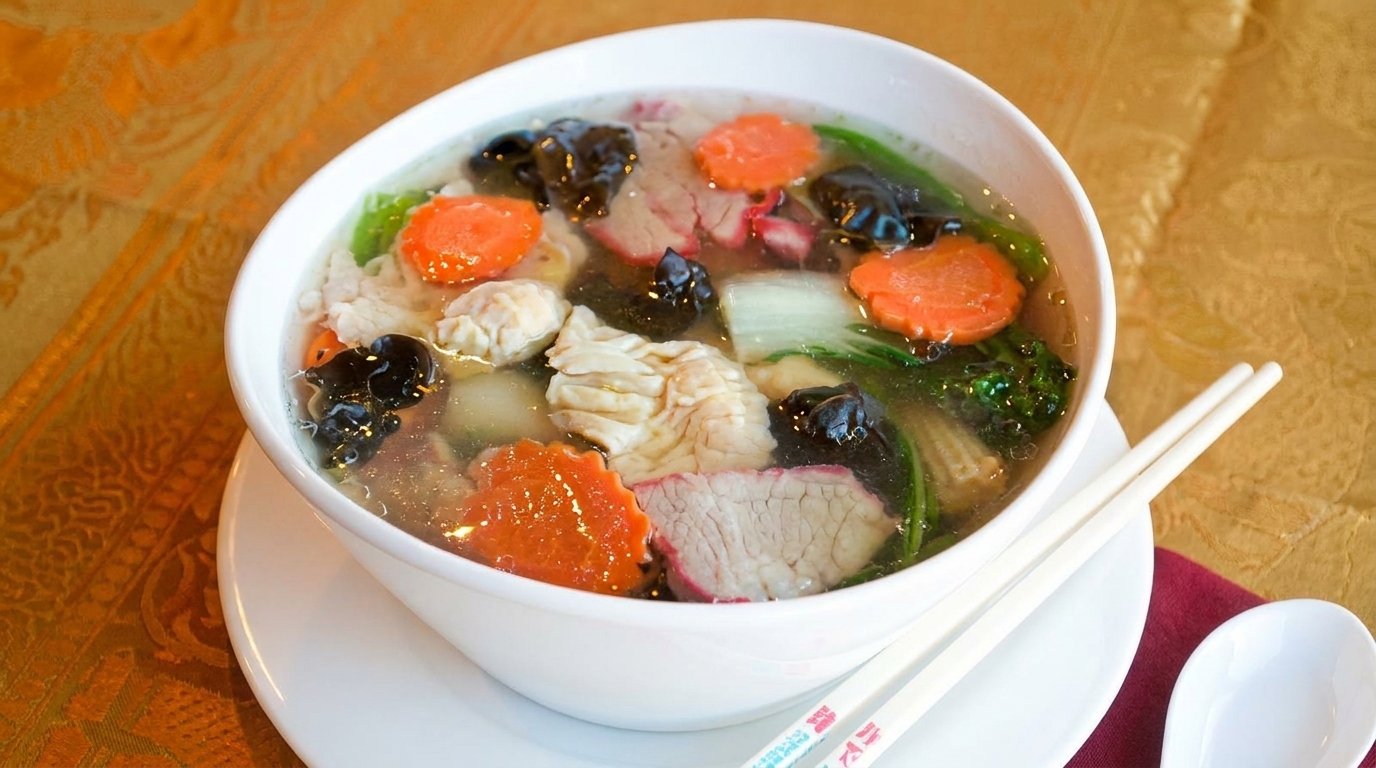 A bowl of clear soup with sliced carrots, bok choy, mushrooms, and various meats, with chopsticks.
