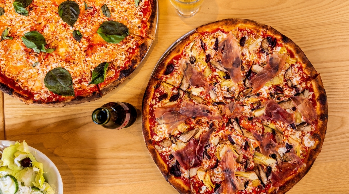 Two pizzas on a wooden table, one with basil and the other with prosciutto, artichokes, and mushrooms.