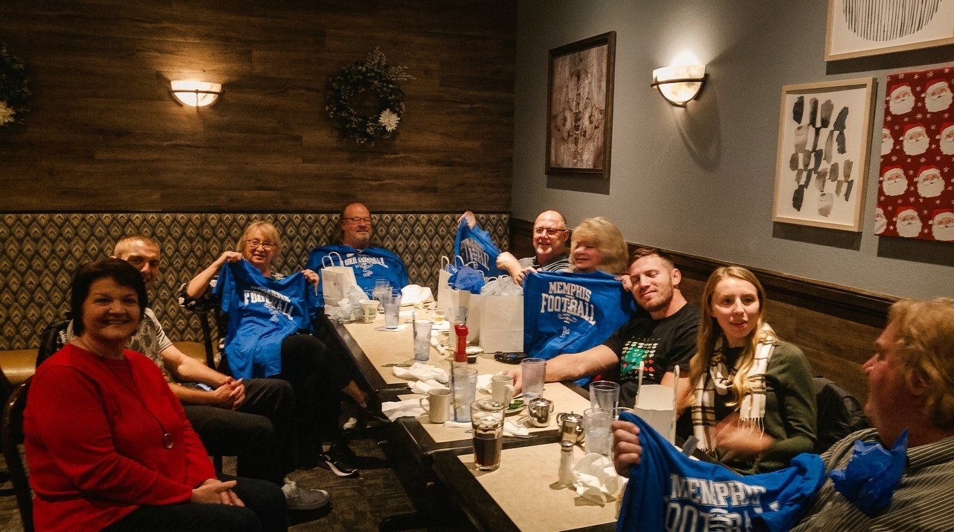 Group of people sitting at a long table in a restaurant, some holding up blue "Memphis Football" shirts.