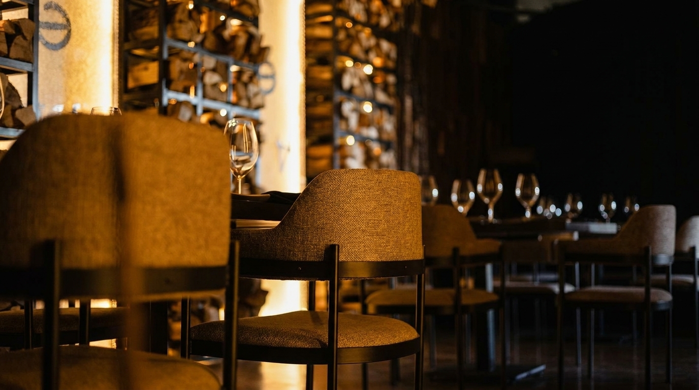 Warmly lit restaurant interior with upholstered chairs, wine glasses, and stacked firewood in the background.