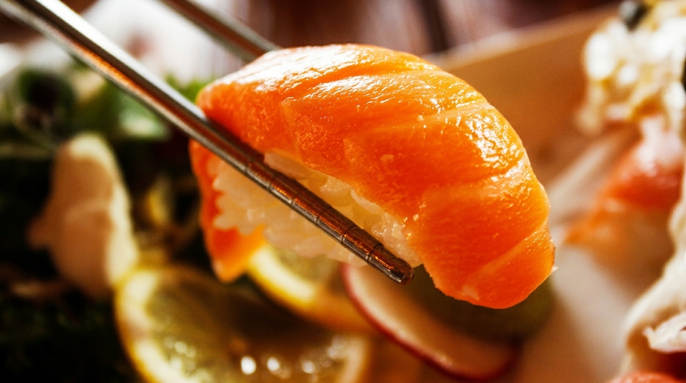 Chopsticks holding a piece of salmon nigiri sushi with rice, against a blurred background.