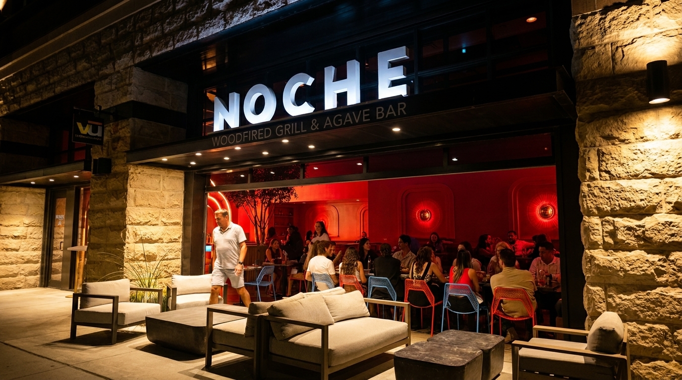 Noche Woodfired Grill & Agave Bar exterior at night, with patrons dining inside and lounge seating outside.