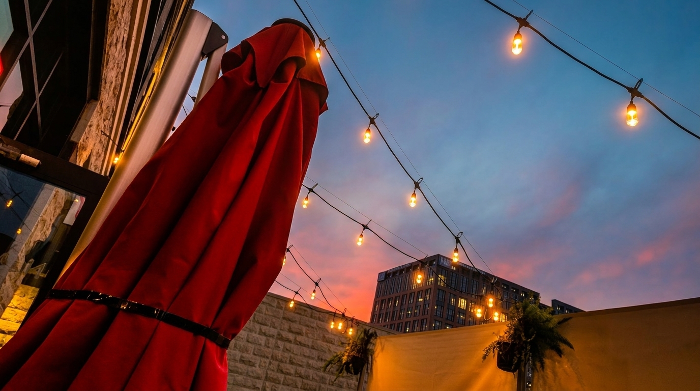 Red patio umbrella and string lights against a vibrant sunset sky with a city building.